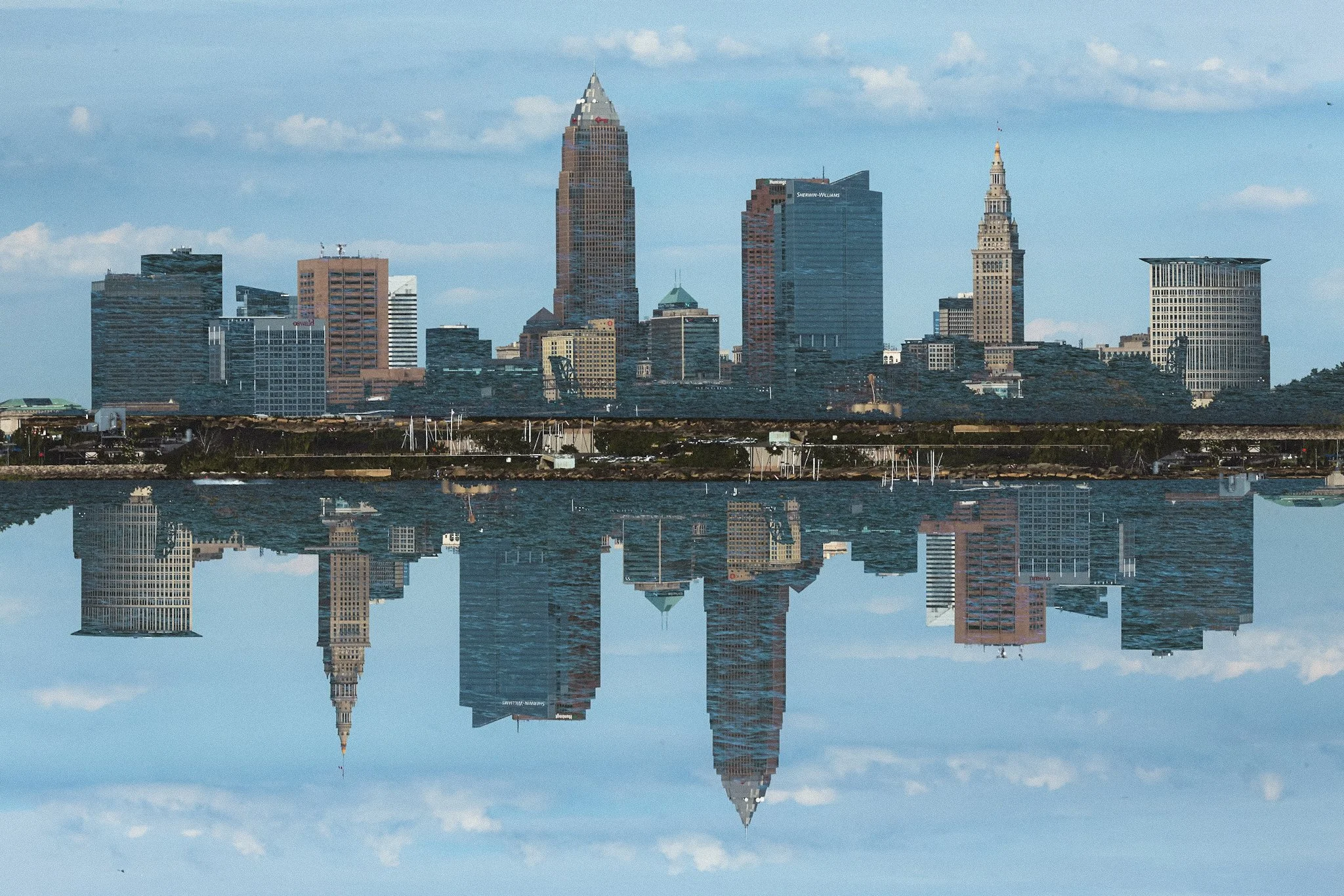 City skyline with tall skyscrapers reflected in a body of water in the foreground, clear blue sky with scattered clouds.