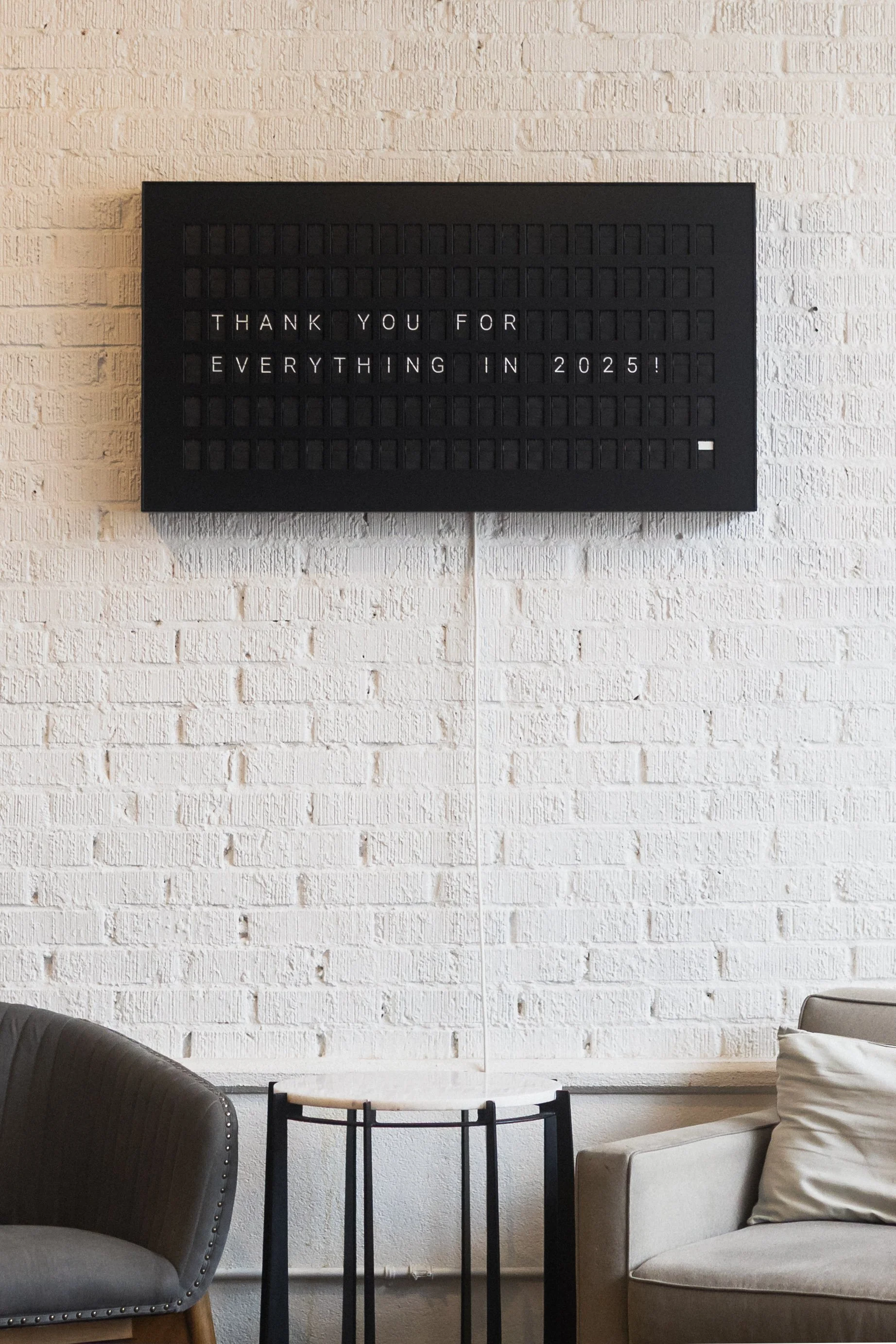 Black letter board mounted on a white painted brick wall reads “THANK YOU FOR EVERYTHING IN 2025.” Below it sits a small round side table between two neutral-colored chairs, creating a minimal, calm interior scene.