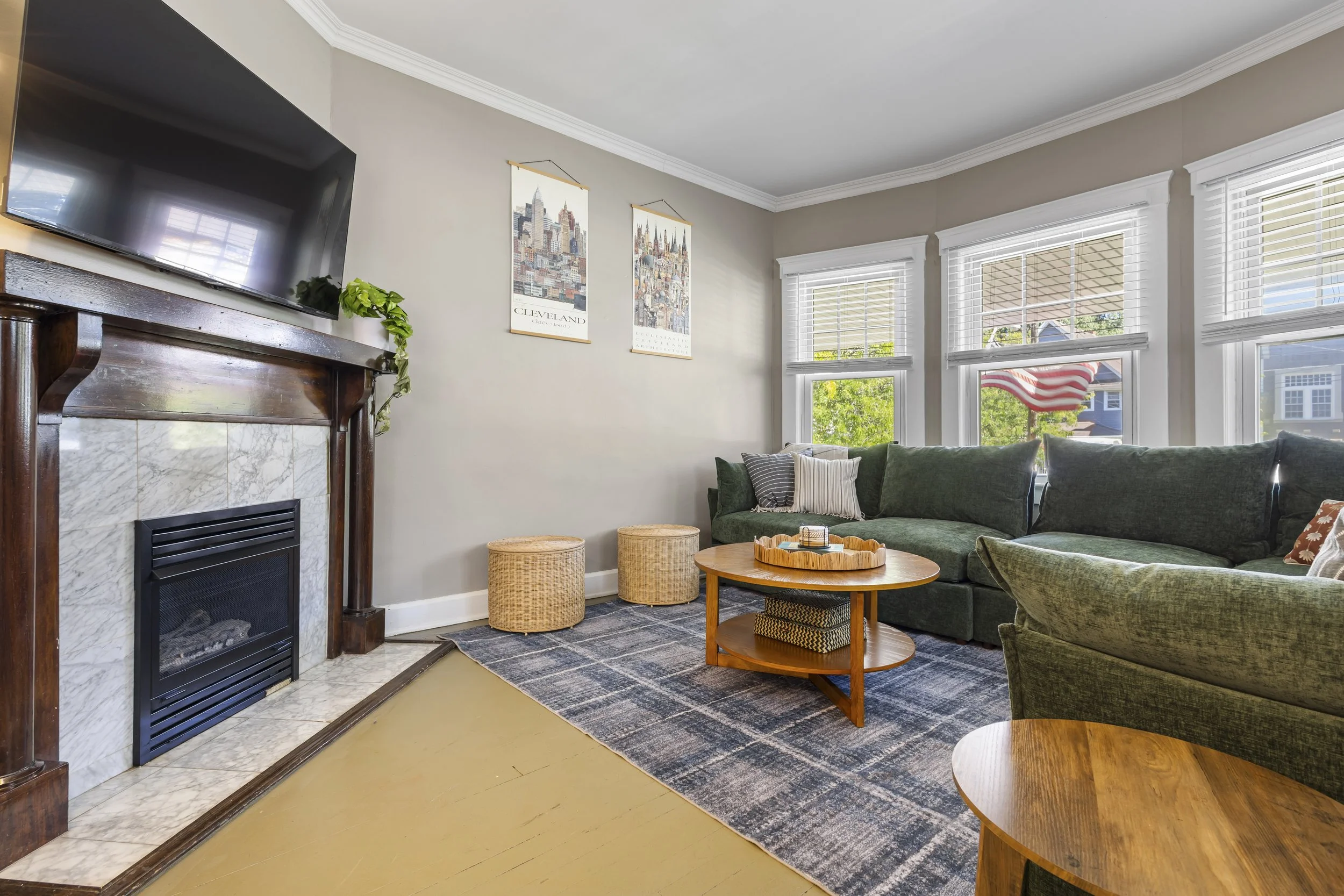 A cozy living room with a fireplace, a large wall-mounted TV, a green velvet sofa with pillows, a round wooden coffee table, wicker stools, and artwork of Cleveland city on the wall. There are large windows with white blinds and an American flag outs