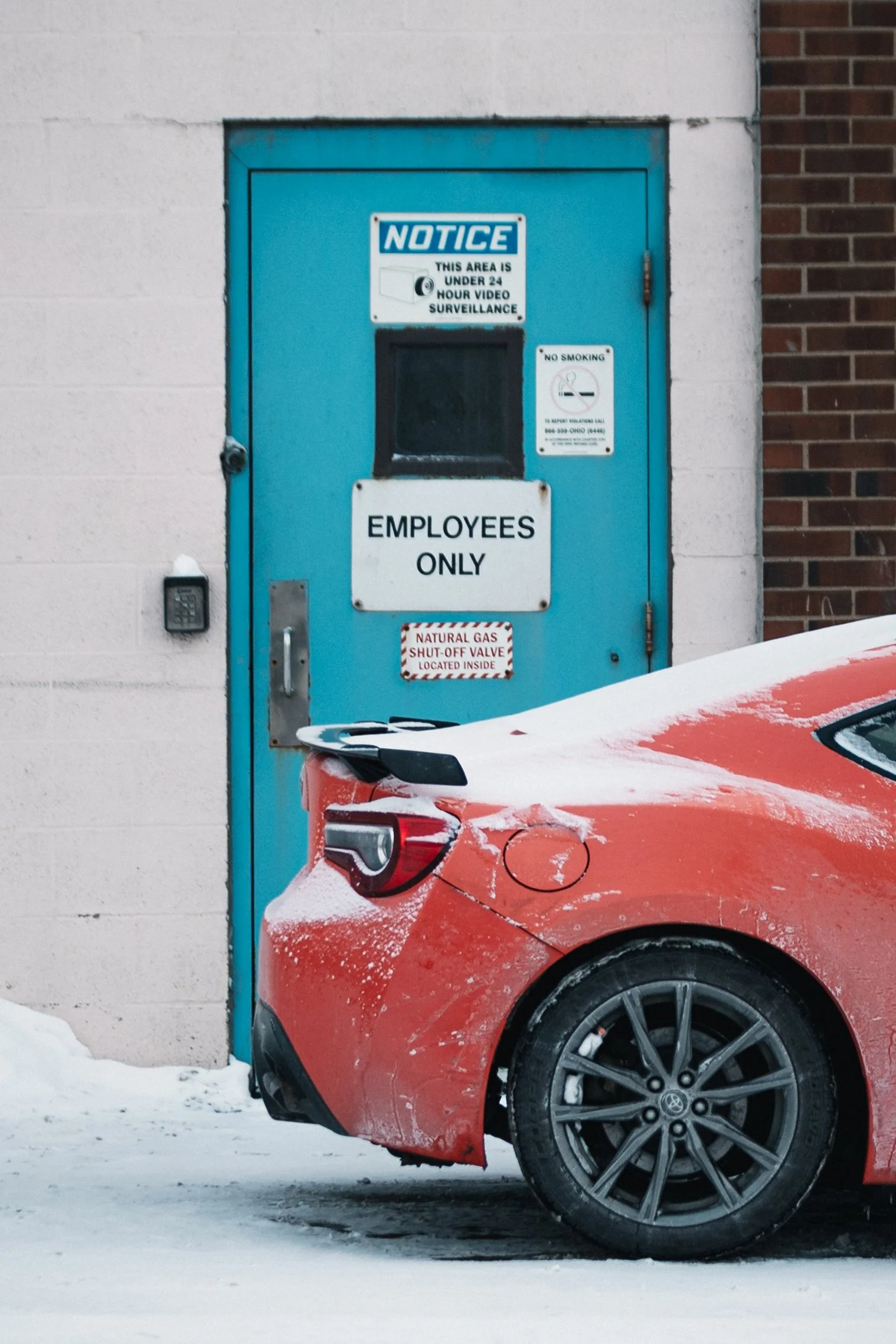 A vibrant red Toyota sports car with snow-dusted roof and hood is parked beside a blue "EMPLOYEES ONLY" door on a brick building wall, under grey skies, with warning signs and a hazard label nearby.