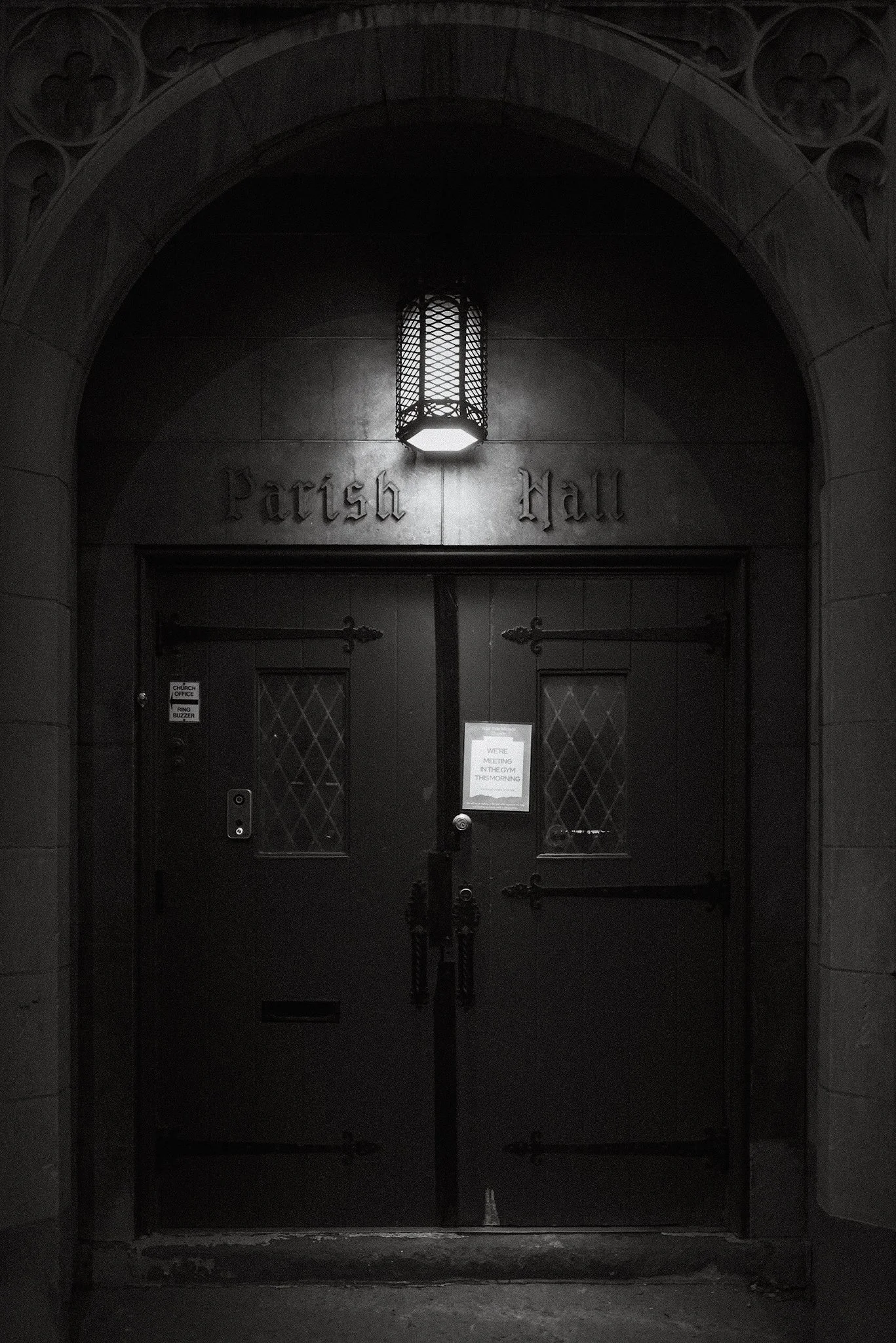 A dark door with a sign that says 'Parish Hall' above it, featuring a hanging lantern light and a small notice on the door.