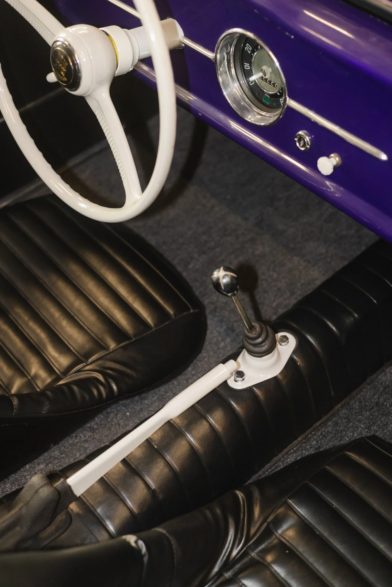 Close-up of a vintage car interior with a purple dashboard, white steering wheel and gauge cluster, black leather bucket seats, and a white manual gear shifter between the seats.