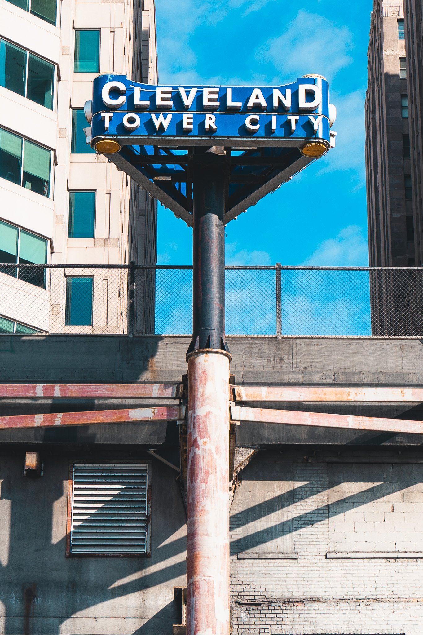 Urban scene featuring a rusted metal girder framework supporting a weathered blue cylindrical structure, possibly a water tank or silo, set against a city backdrop.