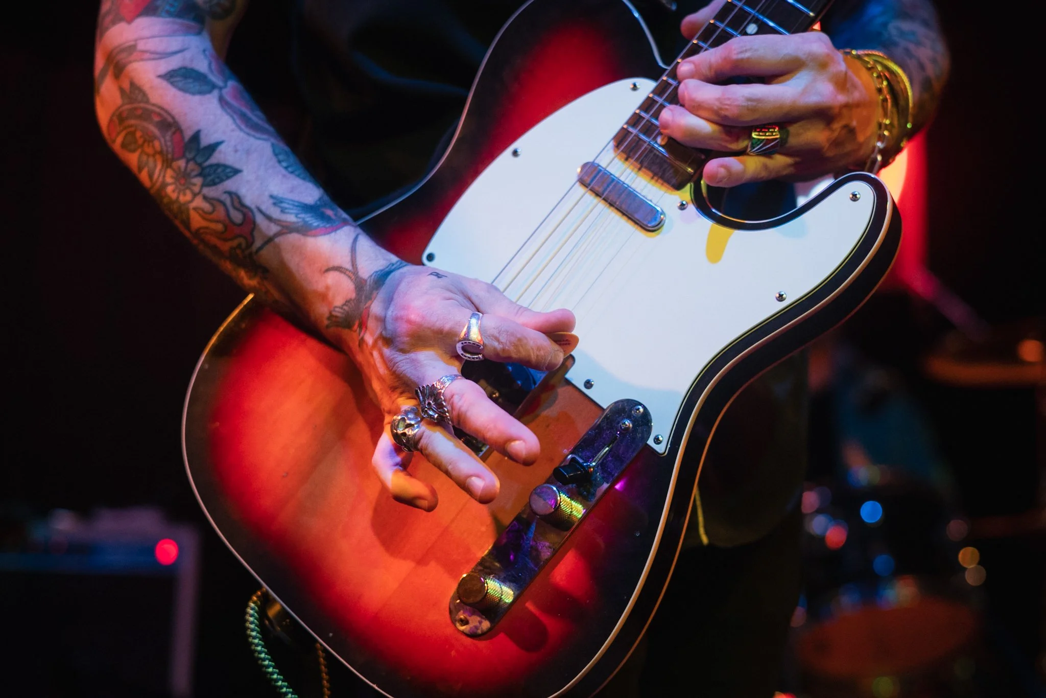 Close-up of tattooed hands with up to three silver rings playing a sunburst Telecaster guitar on stage, highlighted by shallow depth of field and warm bokeh lights.