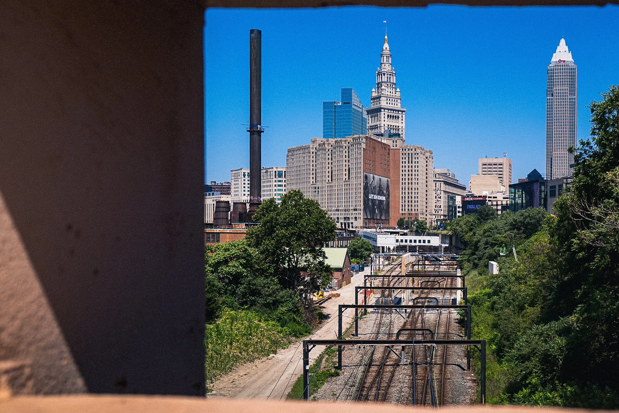 City skyline with tall buildings including the Empire State Building, seen from a railway track with green trees and a clear blue sky in the background.