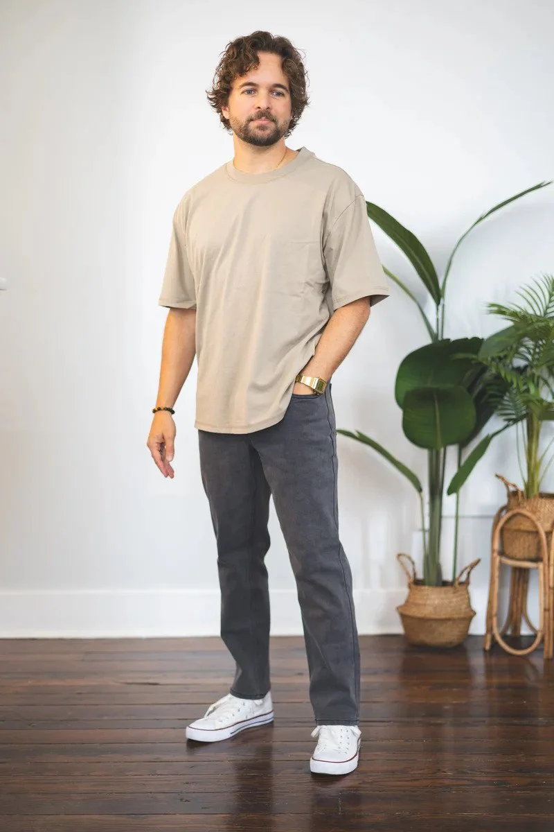 A man with curly hair and a beard standing indoors on wooden flooring, wearing a beige oversized T-shirt, dark jeans, white sneakers, and accessories including a watch and bracelet, with green plants in the background.