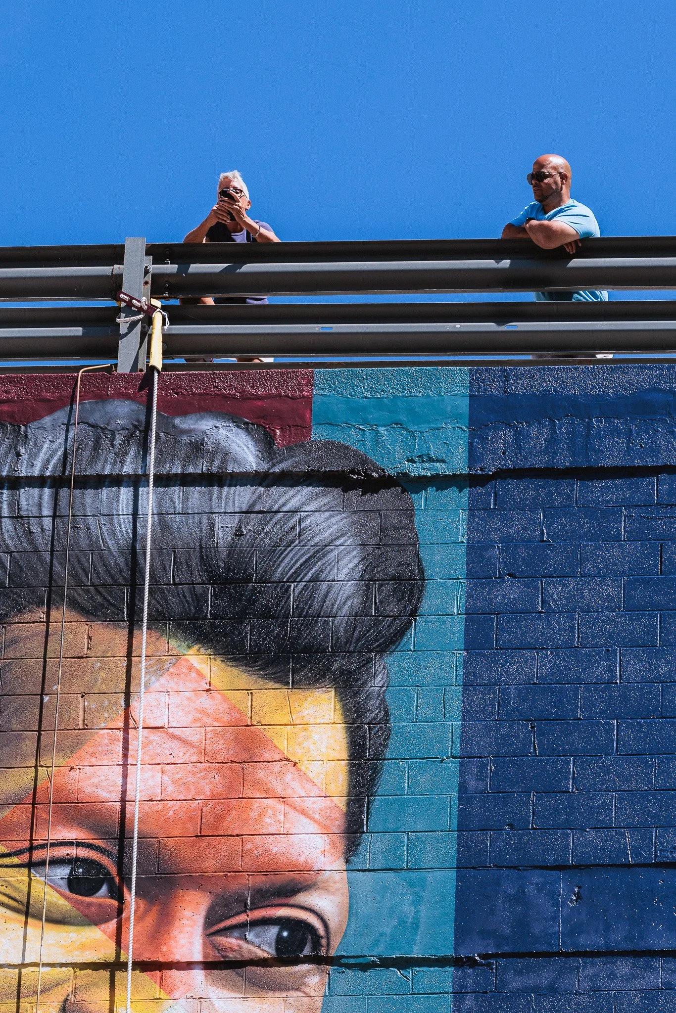 Close-up of a colorful mural of a young child's face painted on a brick wall, with two people standing above it against a clear blue sky.