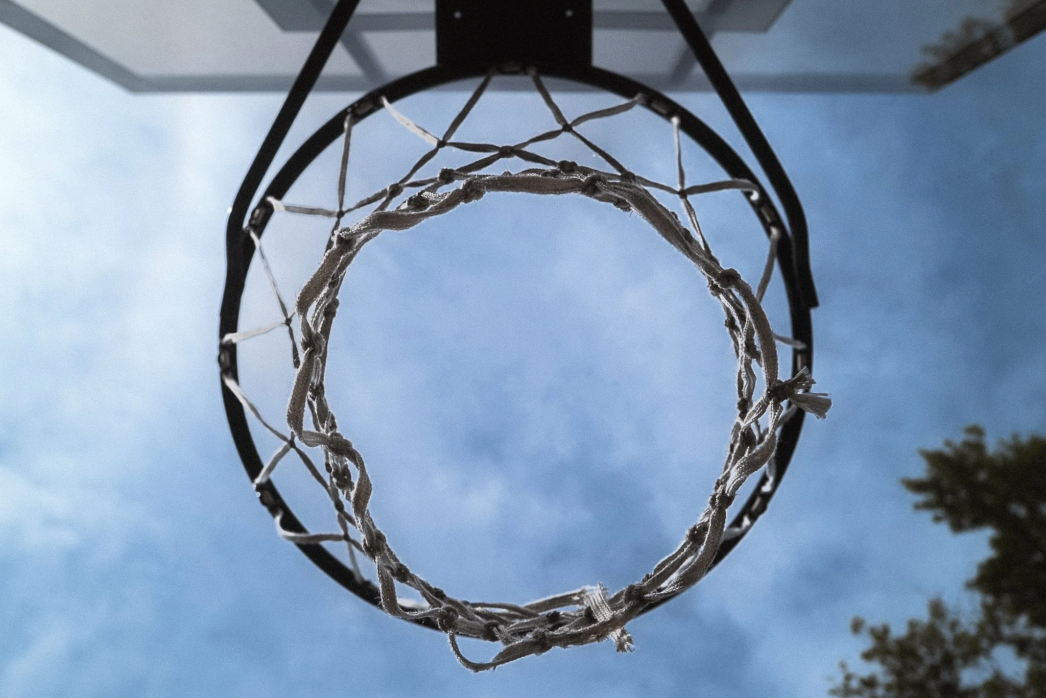 Upward view of a basketball hoop with a net against a blue sky with some clouds and trees in the background.