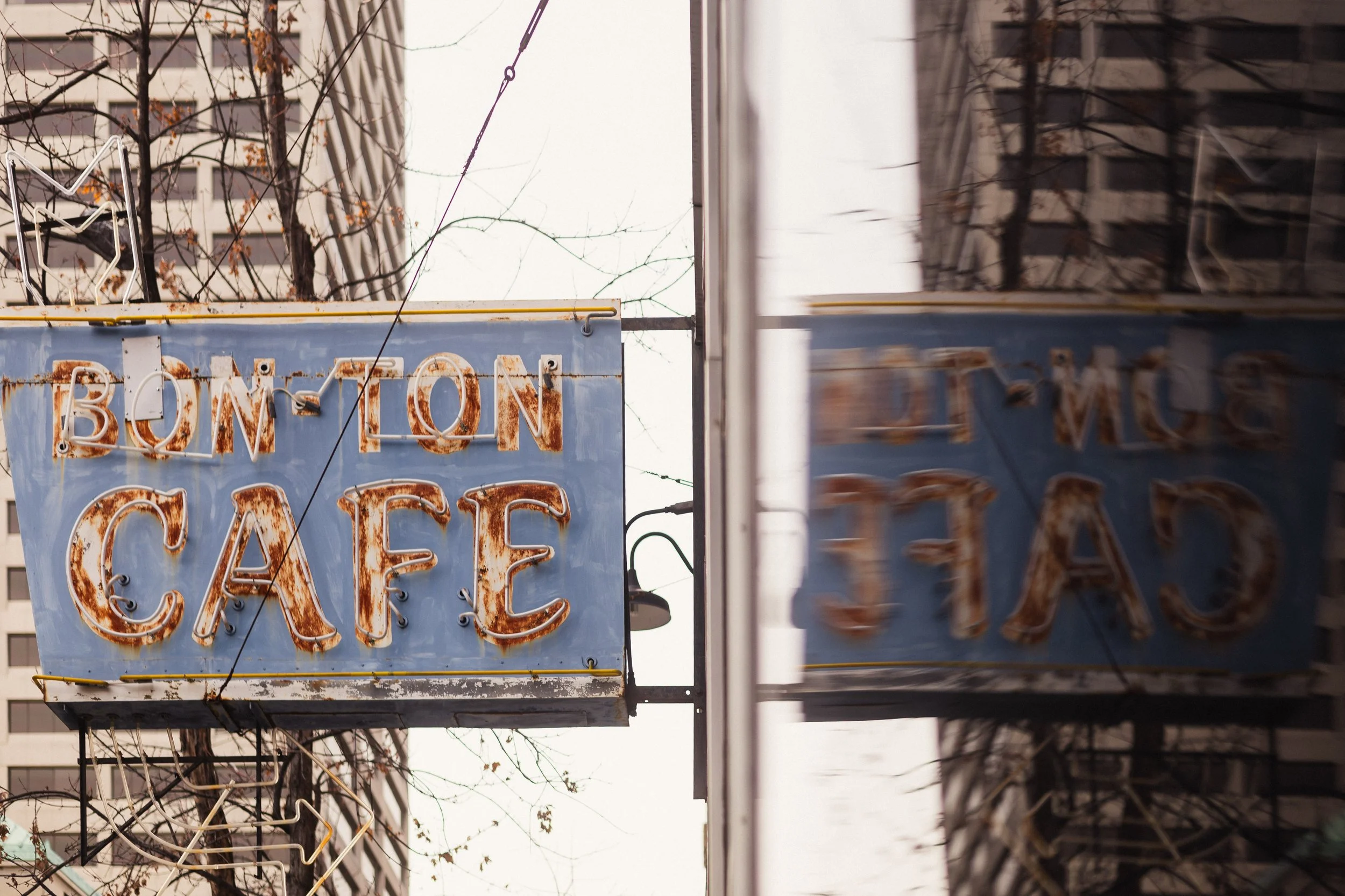 A creative close-up photograph captures a weathered blue neon sign reading "Bon Ton Cafe," suspended from wires amid bare winter tree branches and tall urban buildings under an overcast sky. The composition employs a shallow depth of field and subtle