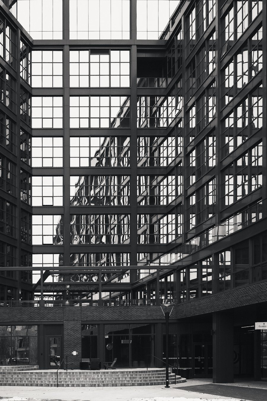 Black and white photograph of a modern building courtyard with a geometric grid of glass windows and steel framing. The strong architectural lines create reflections and symmetry, emphasizing urban minimalism and structure.