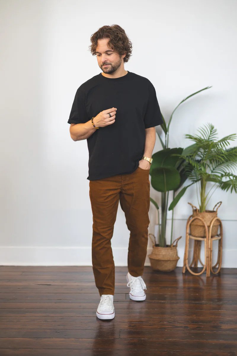 A man with curly brown hair and a beard standing indoors near green houseplants, wearing a black t-shirt, brown pants, white sneakers, and a watch, with his left hand in his pocket and his right hand resting on his chest.
