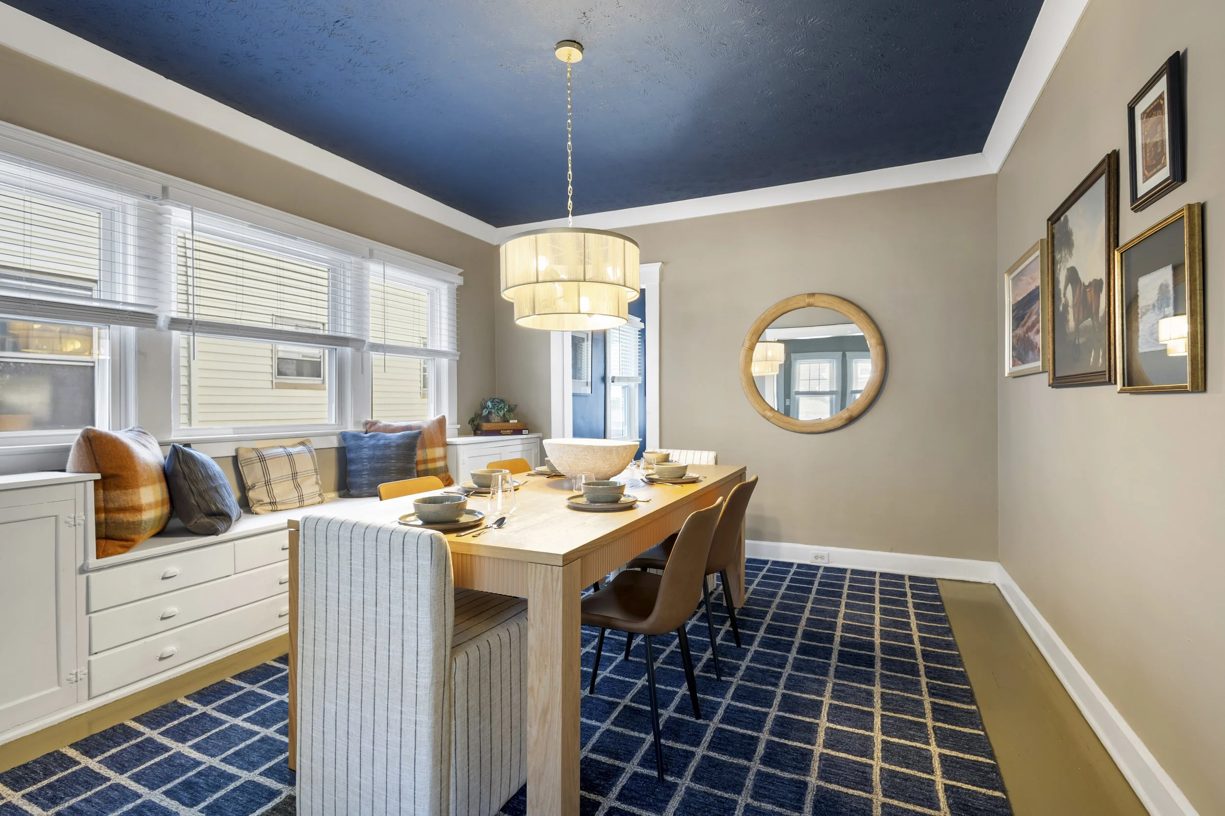 A dining room with a wooden table set with bowls and glasses, surrounded by beige and cream chairs, and a built-in bench with pillows by the window. The room has blue carpet, cream walls, a circular mirror, framed artwork, a white sideboard, and a mo