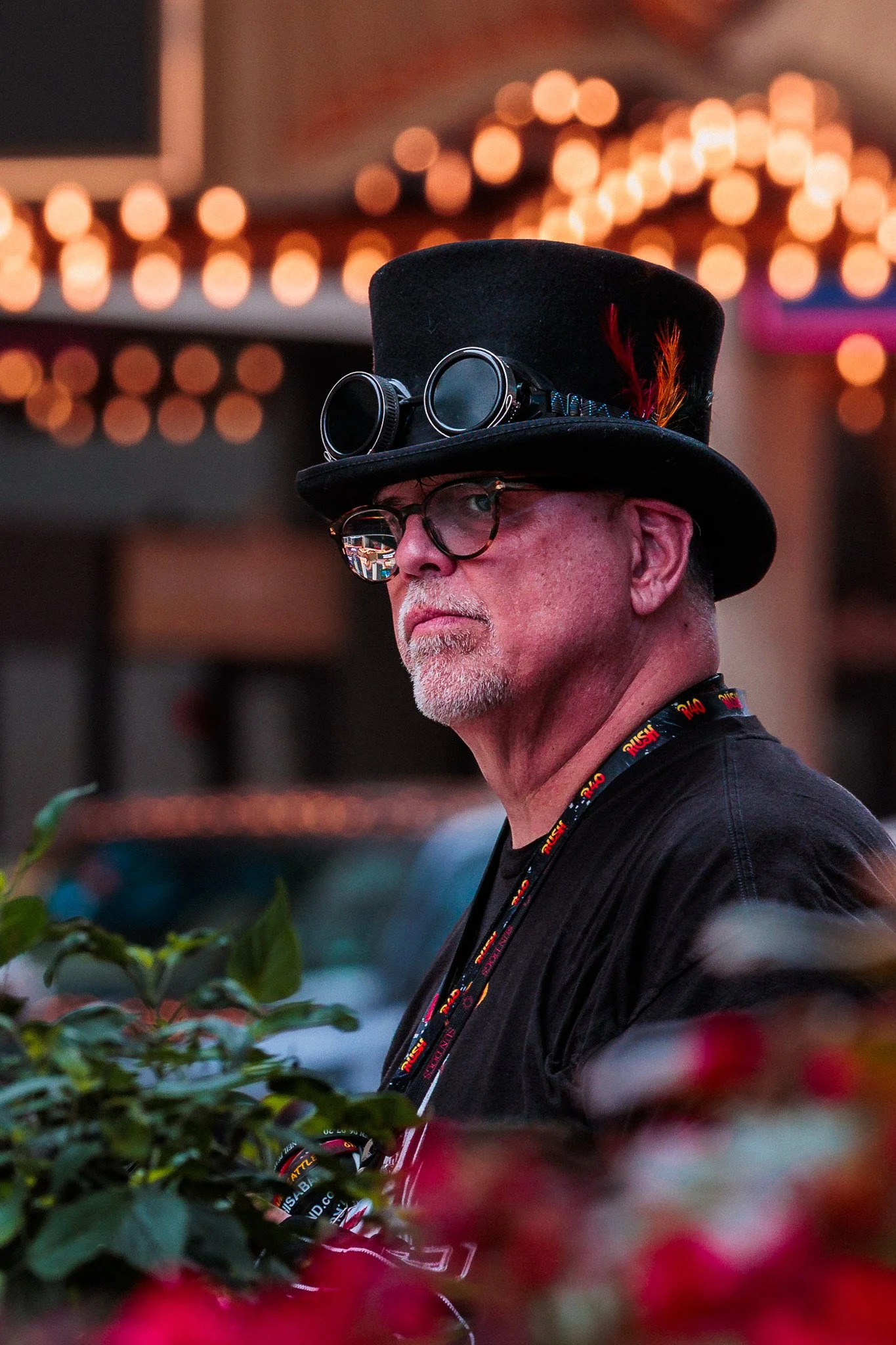 A man with gray hair, glasses, and a beard stands in front of a background with warm, orange string lights. He is wearing a black top hat decorated with goggles and feathers, and a black shirt with a lanyard around his neck.