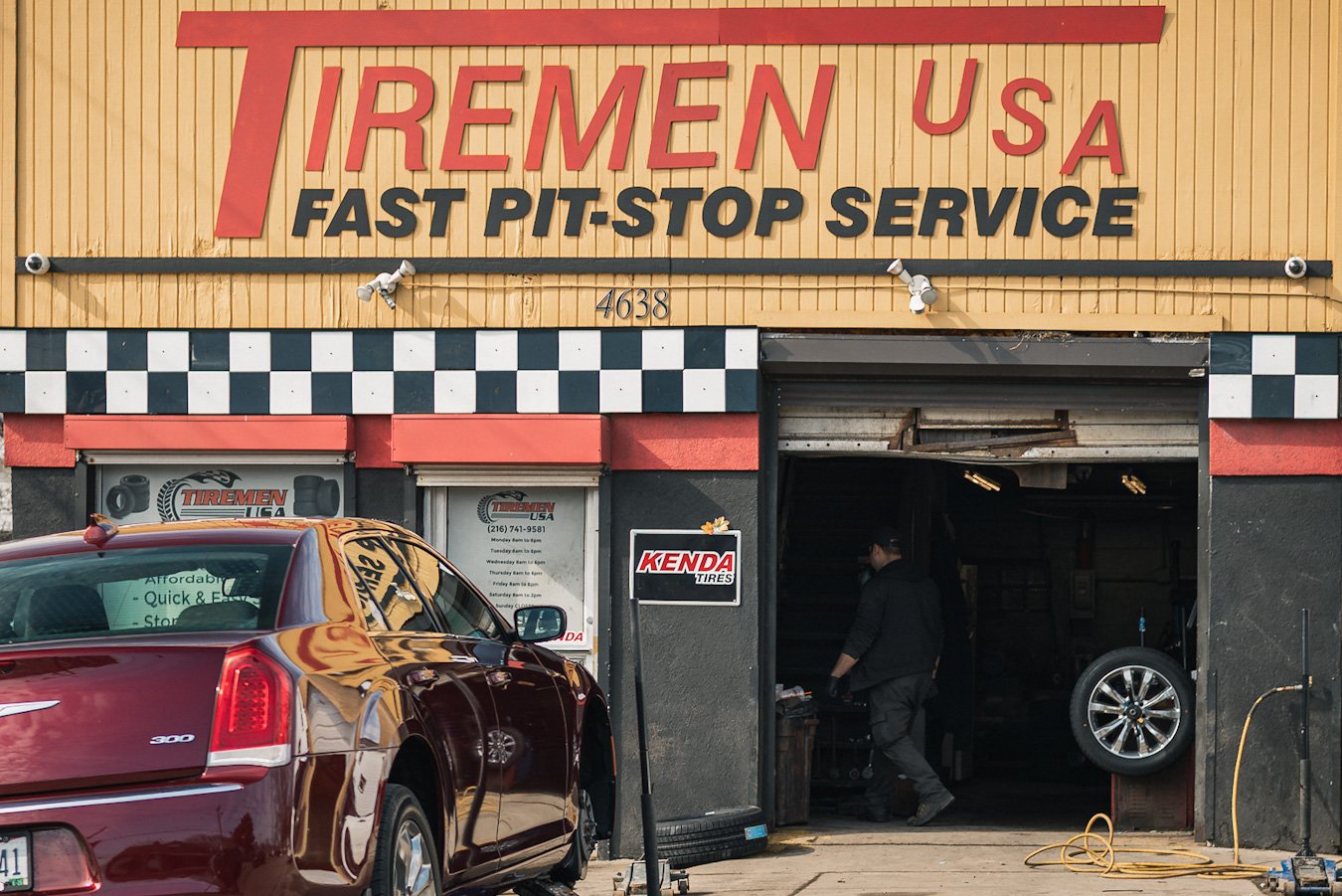A red Chrysler 300 parked outside Tiremen USA, an auto repair shop with a yellow and red facade featuring a bold sign reading “FAST PIT-STOP SERVICE.” A mechanic is visible inside the open garage where a tire is mounted on equipment.