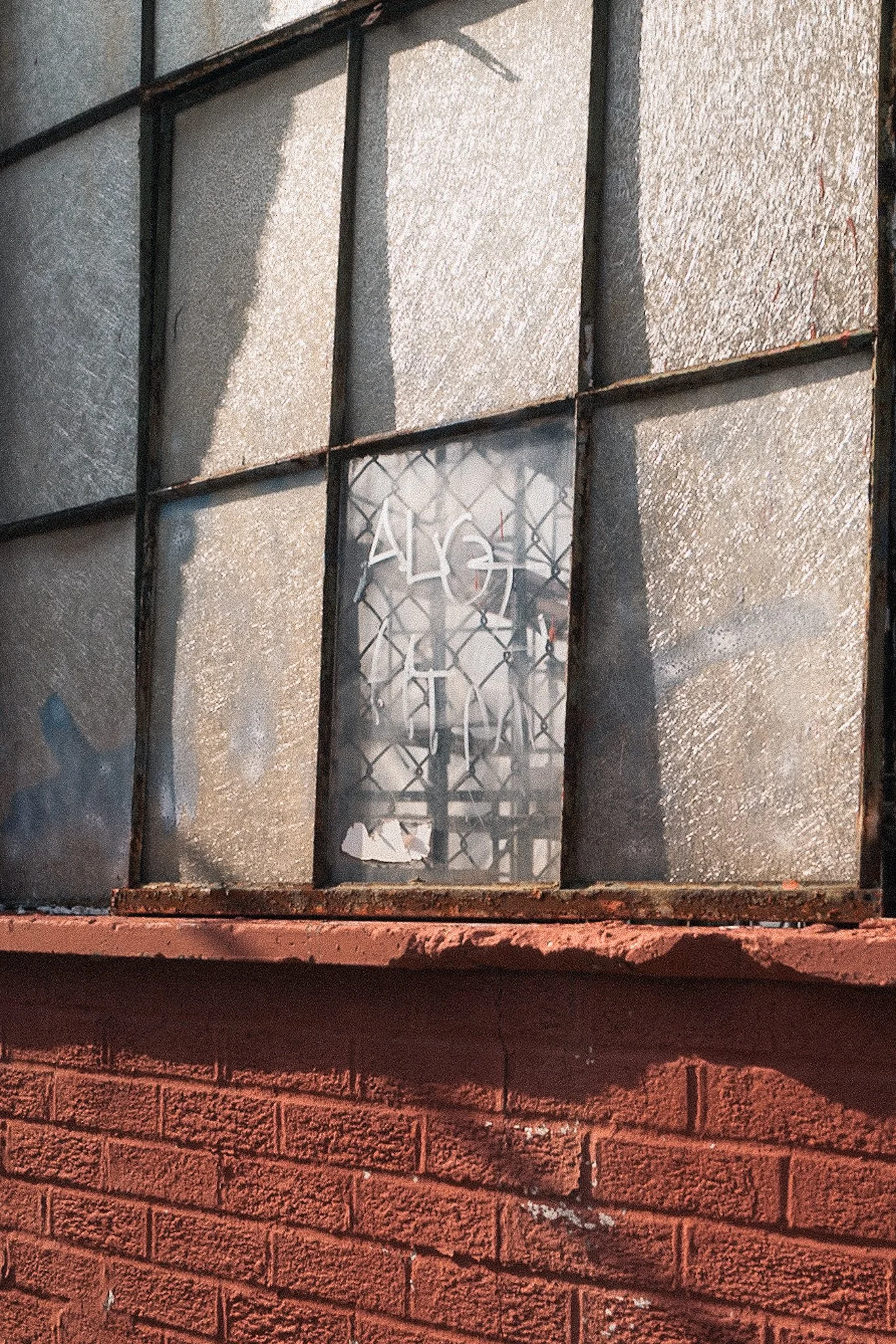 A brick building with a rusty metal window frame and textured glass panes, one of which has graffiti.