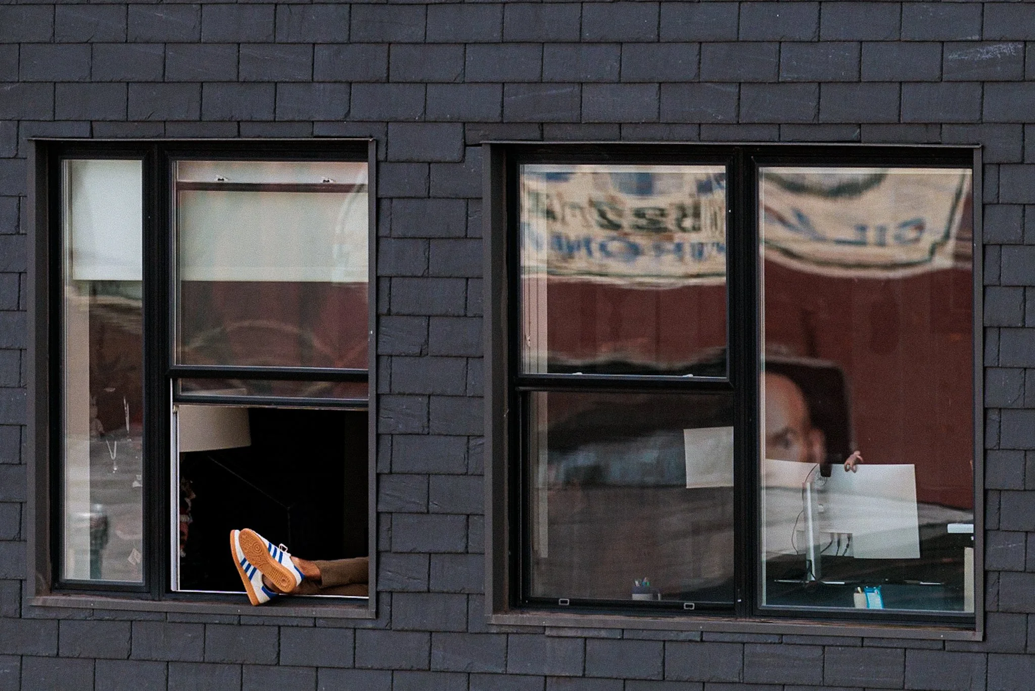 View of a brick building with three large windows. The middle window shows a person sitting inside, their legs extended out wearing blue and white striped shoes. The right window reveals part of an indoor space with furnishings and a reflection of la