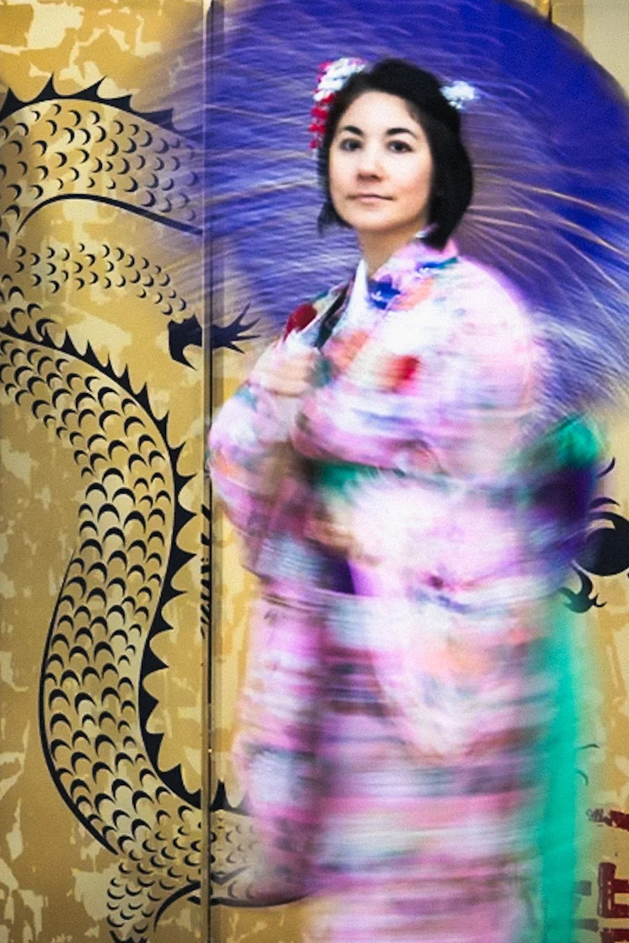 A close-up portrait of an Asian woman in a colorful floral kimono posing gracefully with a large blue parasol held over her shoulder, her hair adorned with pink flowers, against a tall beige folding screen painted with a black Chinese dragon design, 