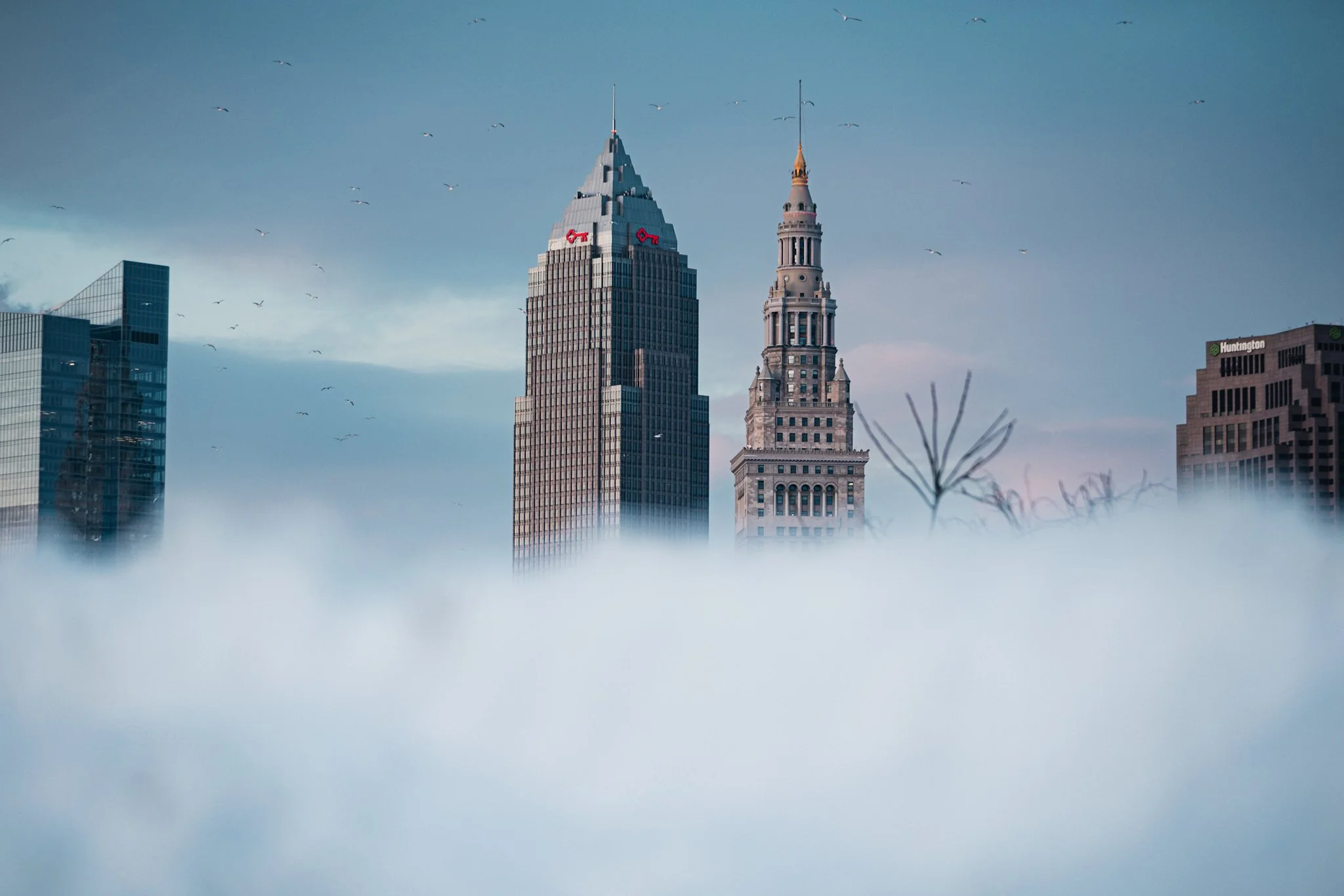 A dramatic aerial view of Cleveland's downtown skyline featuring the iconic Key Tower with its red "Key" logo and pyramid spire flanked by the historic Terminal Tower's clock tower, other modern skyscrapers, bare winter trees, and a thick layer of lo