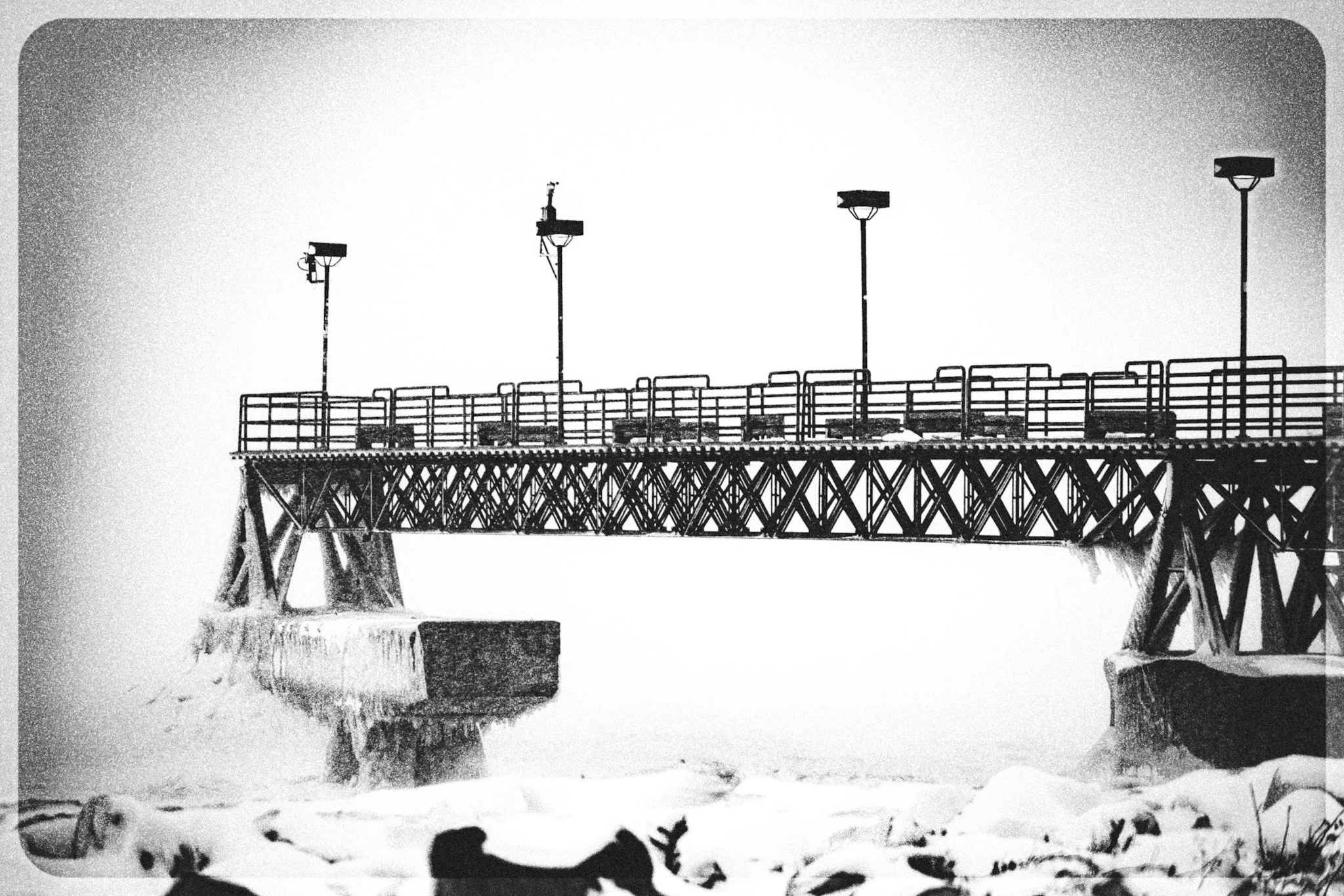 A metal pier on Lake Erie disappears into a whiteout snow squall, its railings and supports coated in ice while waves and snow blur the frozen shoreline in the foreground.