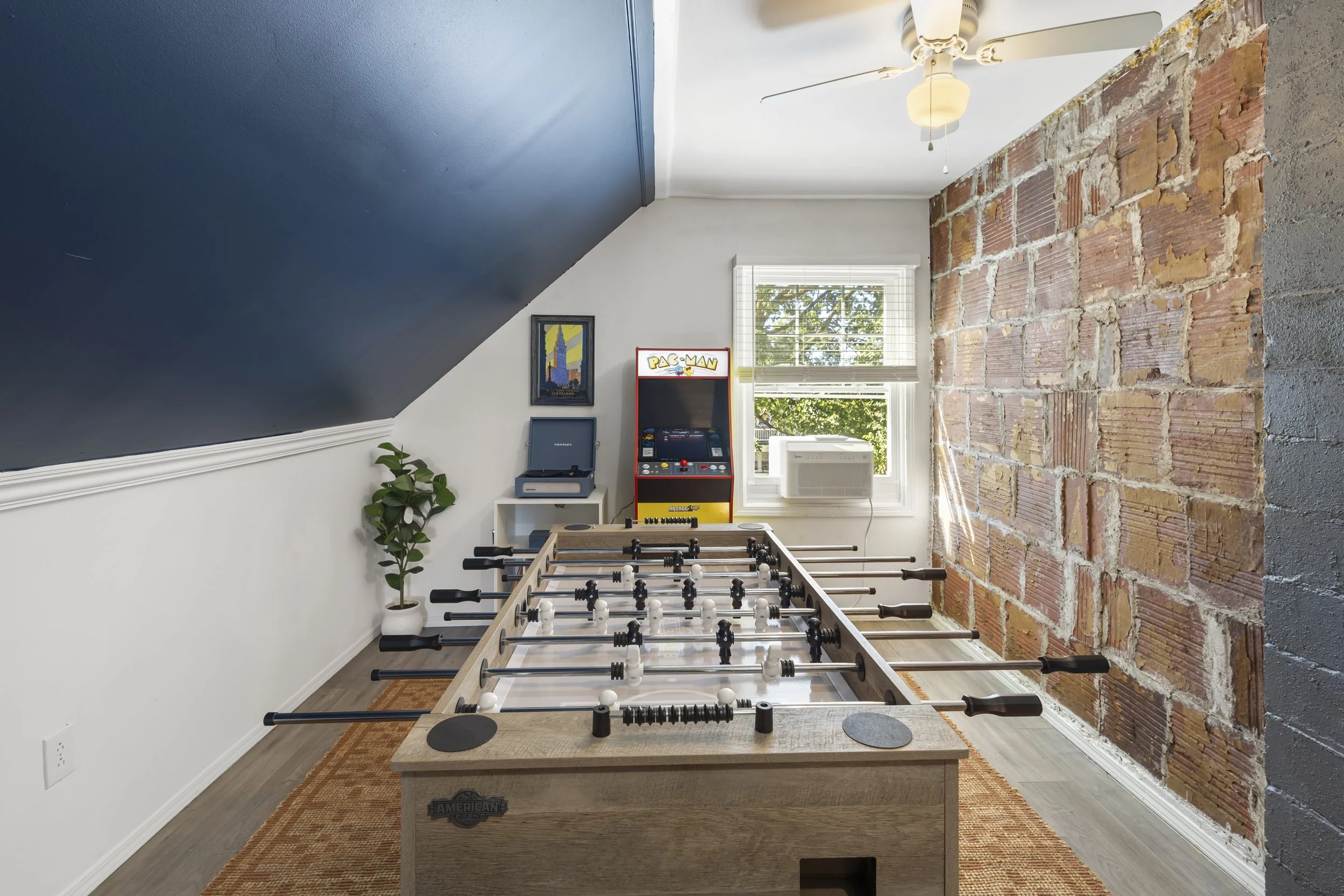 A room with a foosball table, arcade game, and artwork, featuring a brick accent wall, a large window, and a ceiling fan.