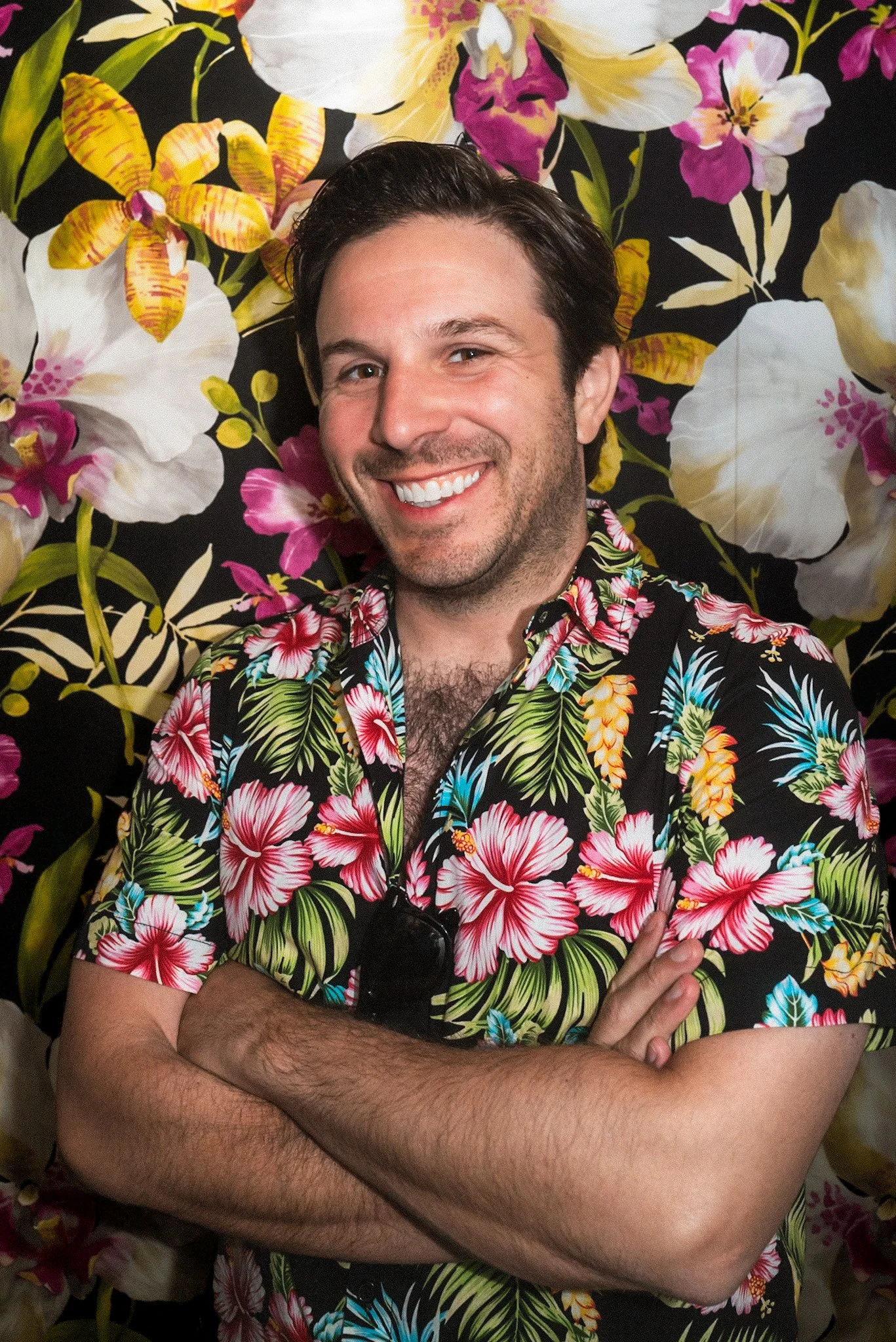 A man with dark hair and a beard smiling, wearing a colorful Hawaiian shirt, standing in front of a floral background with large white, pink, and yellow flowers.