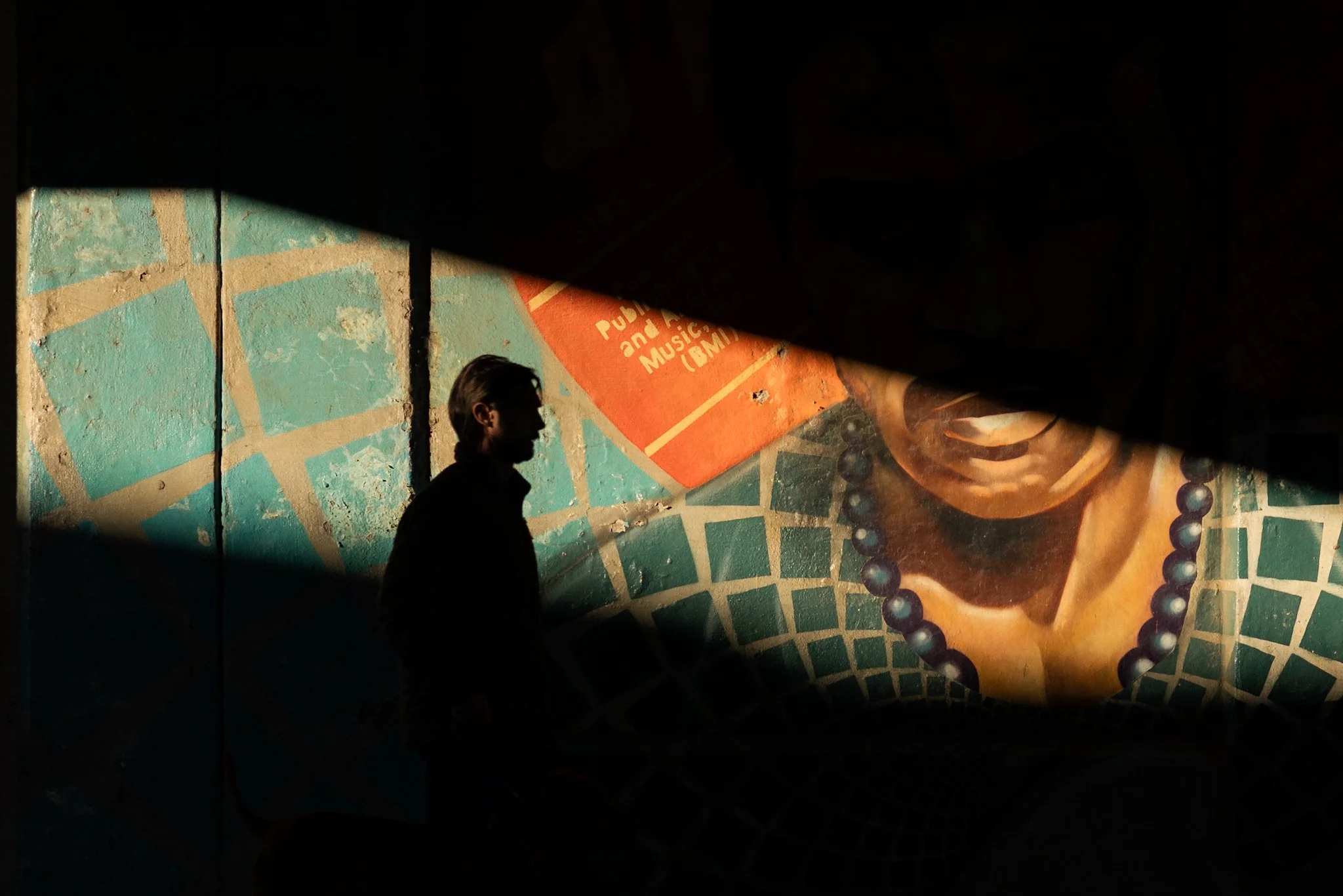 A close-up street photograph captures a man with long hair viewed from the side, standing on a shadowed urban sidewalk. The mural has a green and blue pattern with orange accents. Dramatic golden sunlight streams from the upper right, creating sharp 