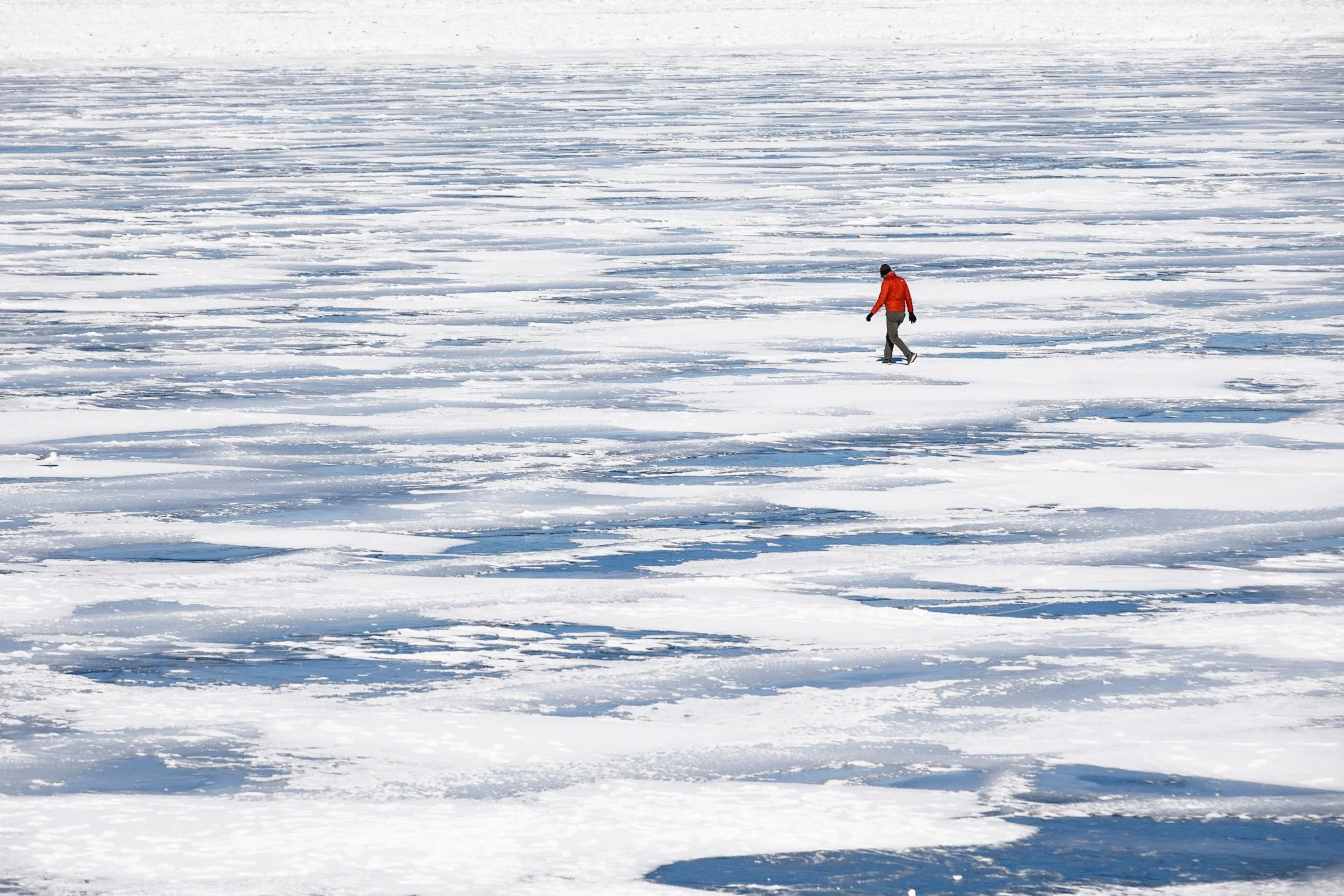 A person wearing a red jacket walks across the frozen surface of Lake Erie in Cleveland, Ohio, surrounded by patches of snow and ice stretching to the horizon.