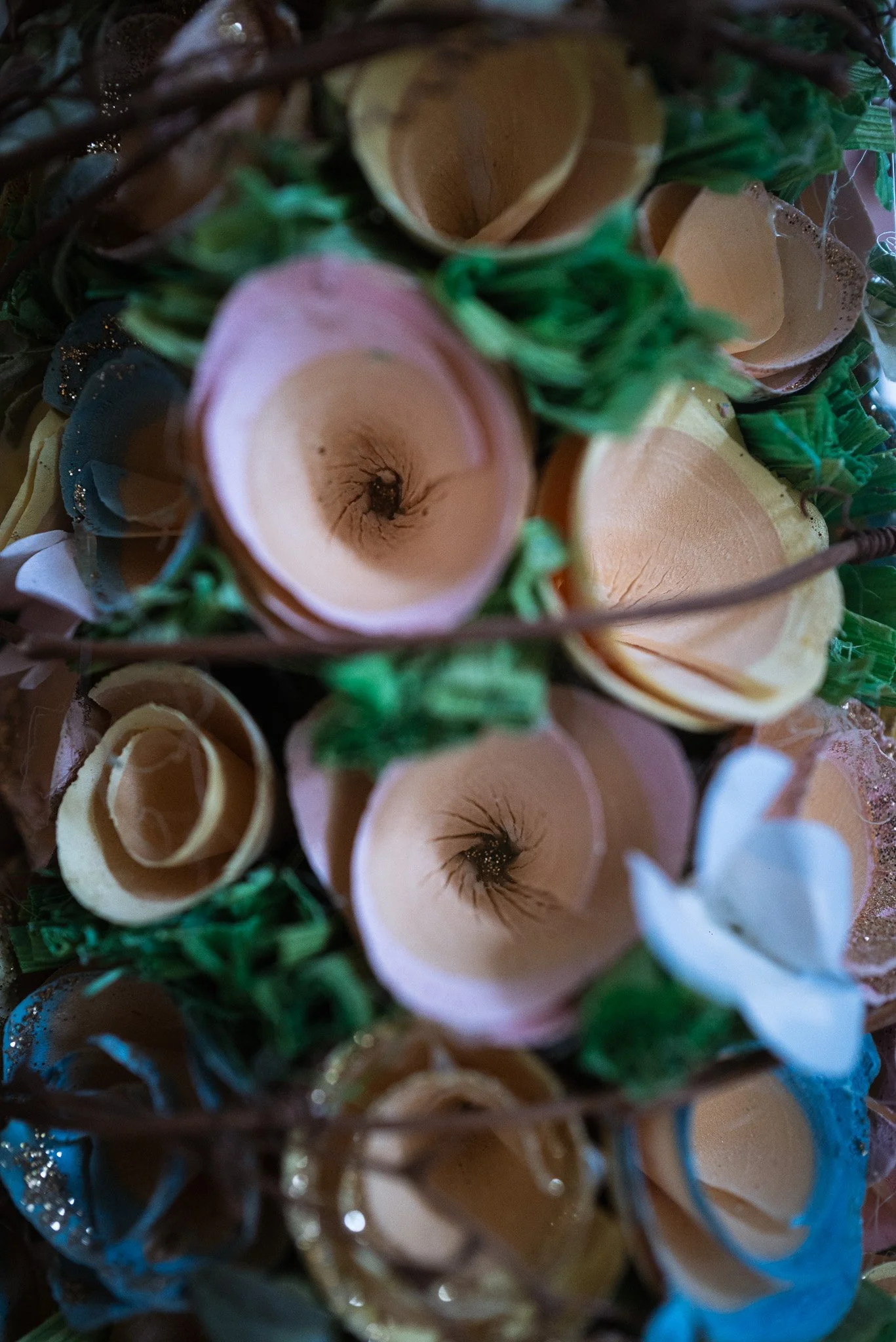 A close-up view of a basket filled with artificial flowers made of rolled and shaped paper or fabric, resembling roses and other blooms, with green decorative foliage woven throughout.