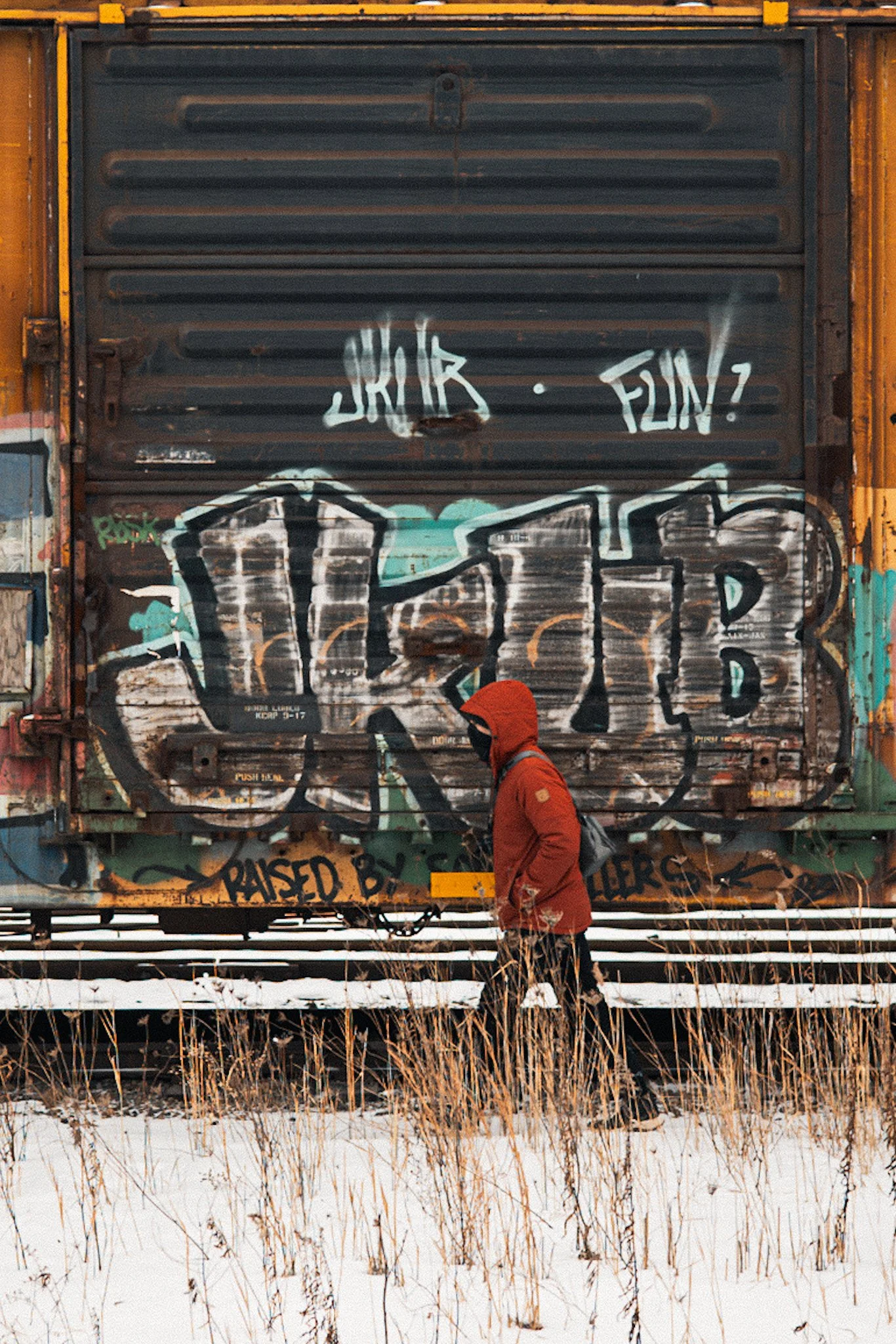A person in an orange hooded parka walks through a snowy train yard in front of a weathered freight car covered in colorful graffiti and rust.
