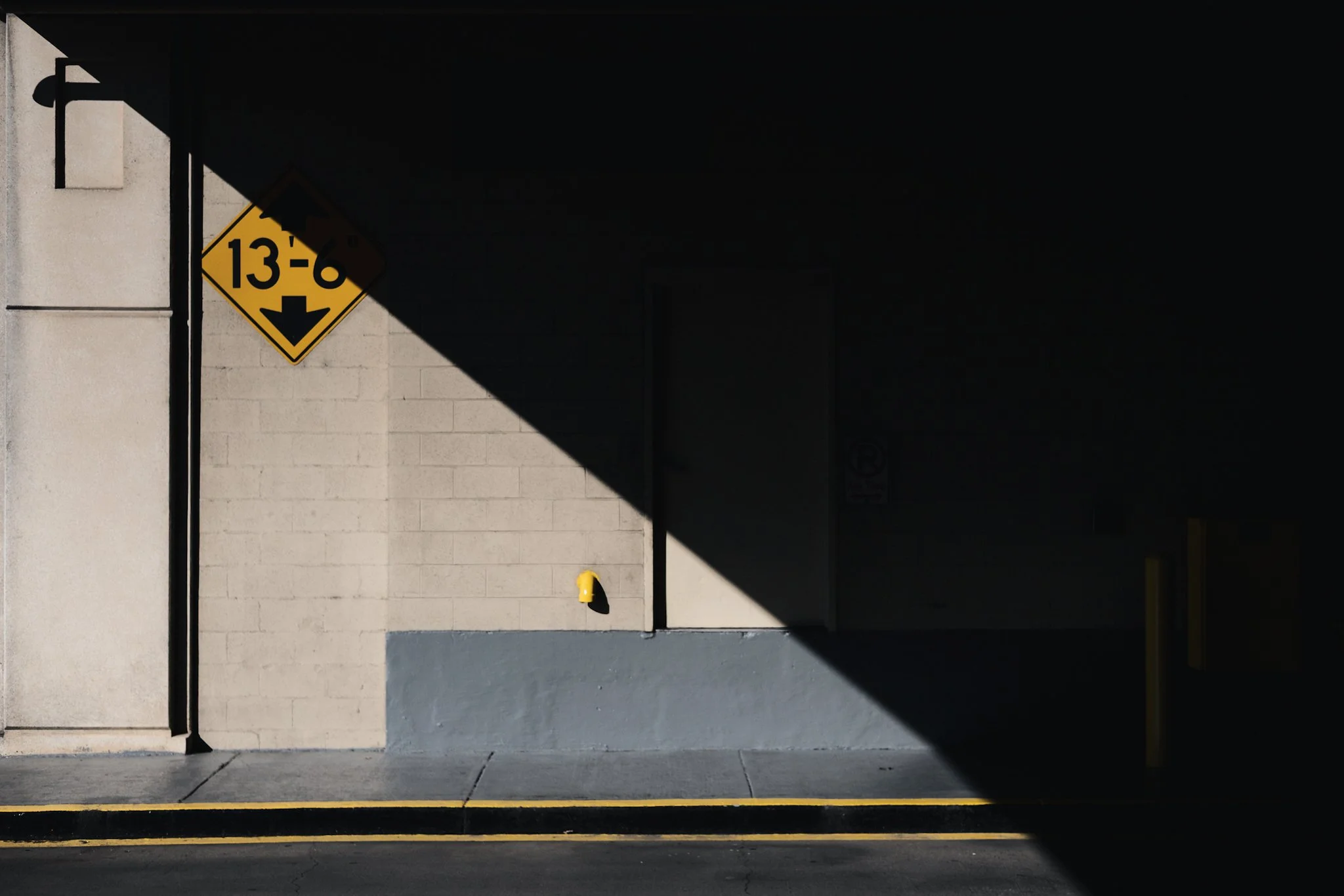 A concrete wall partially illuminated by sunlight, featuring a yellow clearance sign reading "13'-6”" and a sharp diagonal shadow dividing the frame into bright and dark halves.