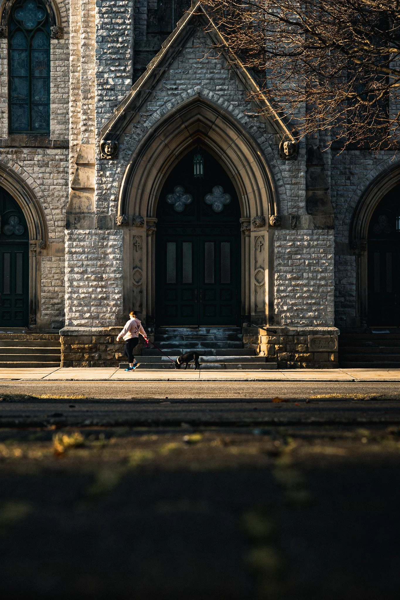 Person walking a dog past a stone church facade, bathed in warm golden hour light with long shadows stretching across the sidewalk.