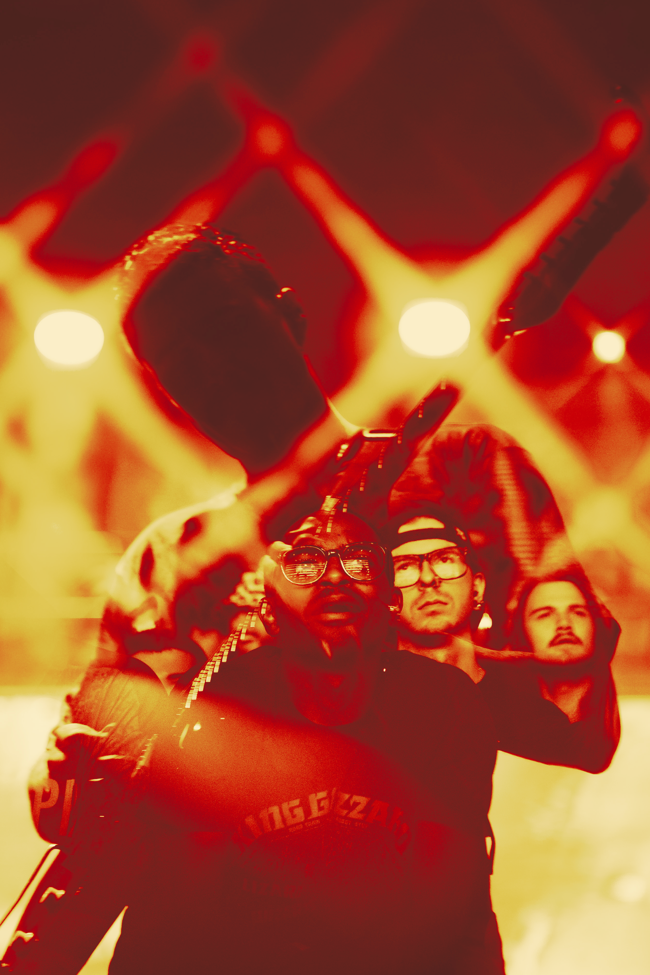 Audience at a concert under red lighting, some wearing glasses and band merchandise, looking up towards the stage with expressions of enjoyment.