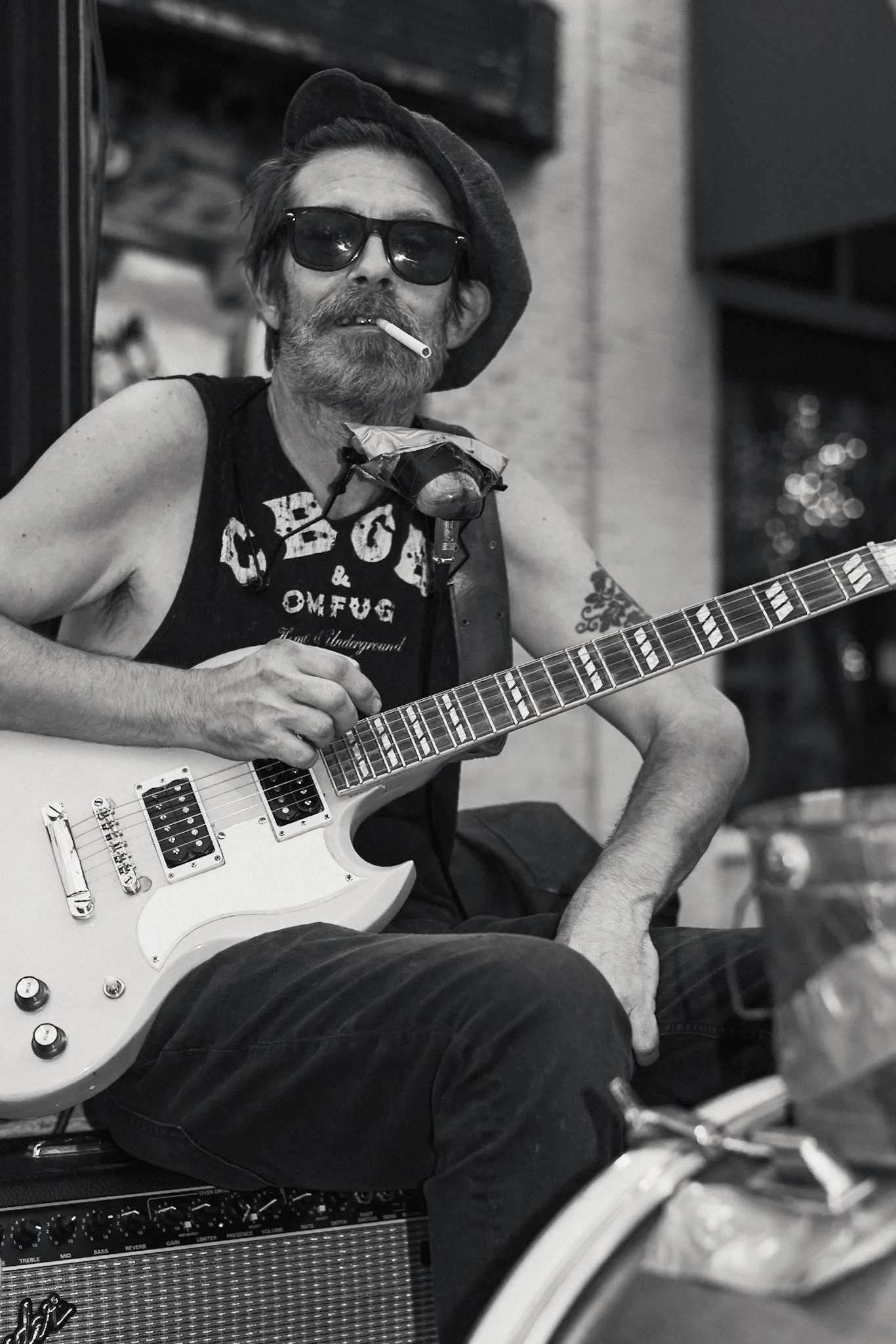 Black and white photo of a street musician sitting outdoors with an electric guitar on his lap. He wears sunglasses, a beret, and a sleeveless CBGB T-shirt, with a cigarette hanging from his mouth. A harmonica rests in a holder around his neck, and h