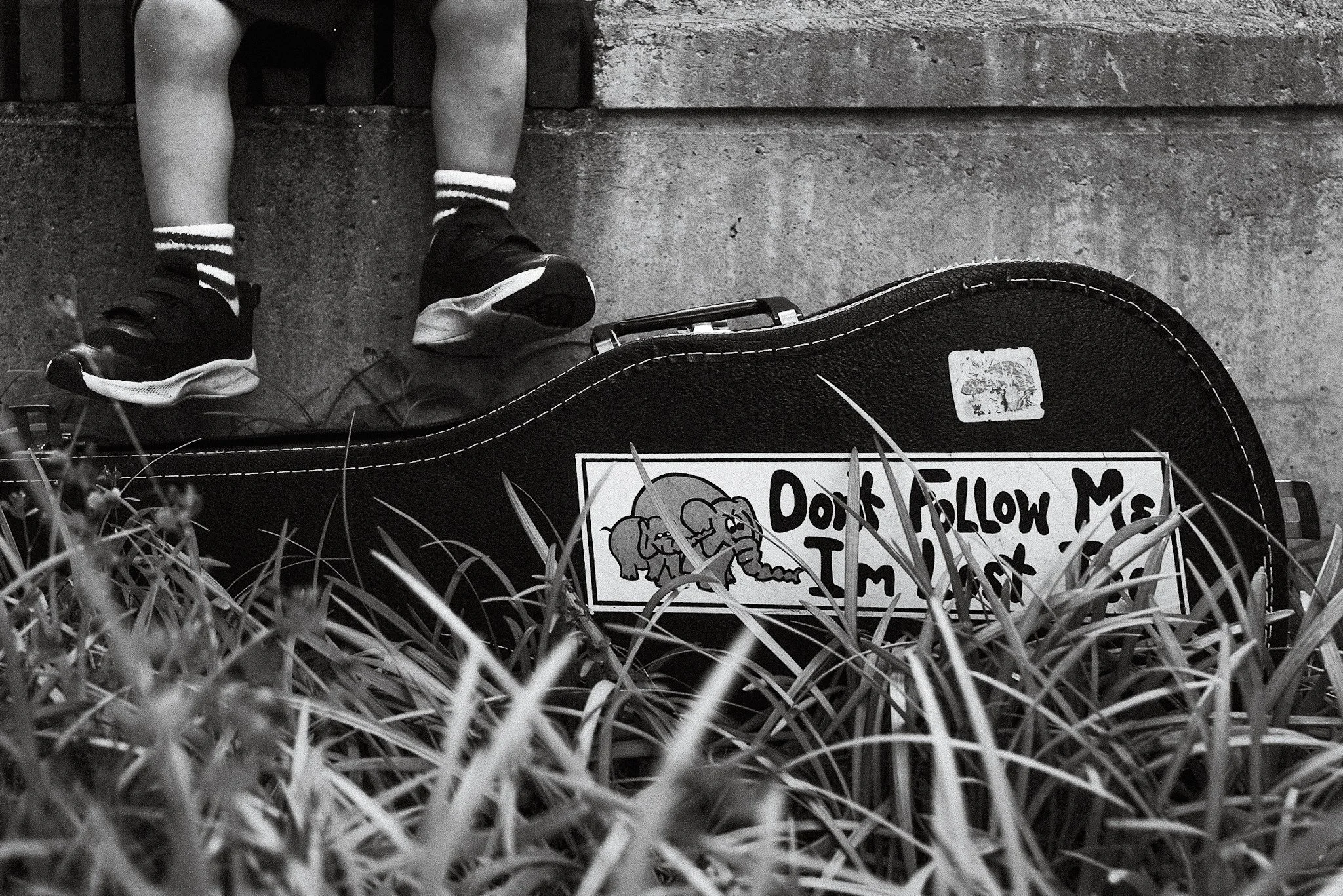 Close-up black-and-white photo of a child's legs, wearing striped socks and sneakers, sitting on a concrete ledge. In front, there is a sign with cartoon elephants and partially visible text that reads 'Don’t Follow Me' amid grass.