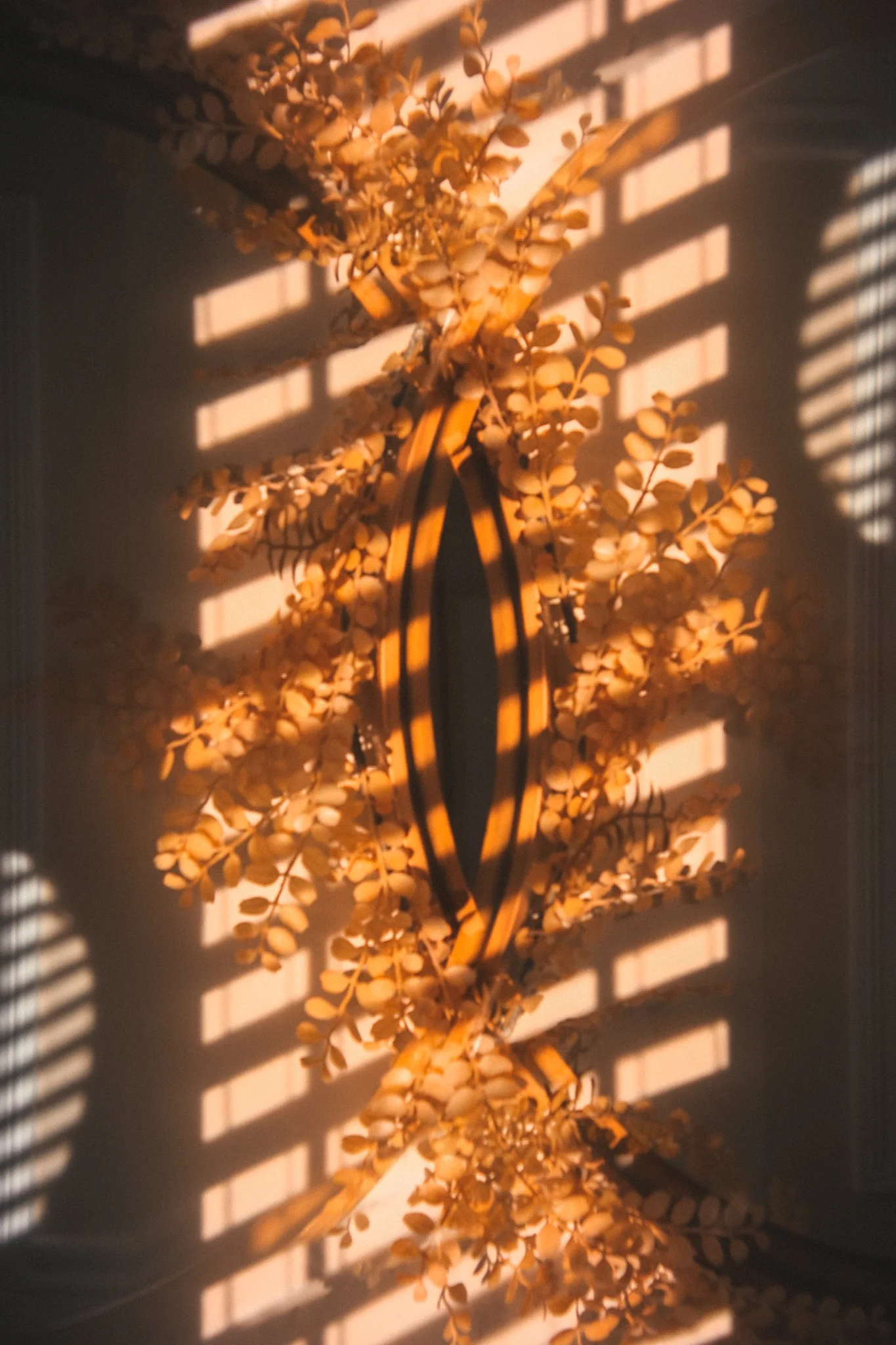 A warm double exposure image featuring overlapping fall leaves and striped shadows from window blinds, forming an oval silhouette in glowing orange and golden tones against a softly lit background