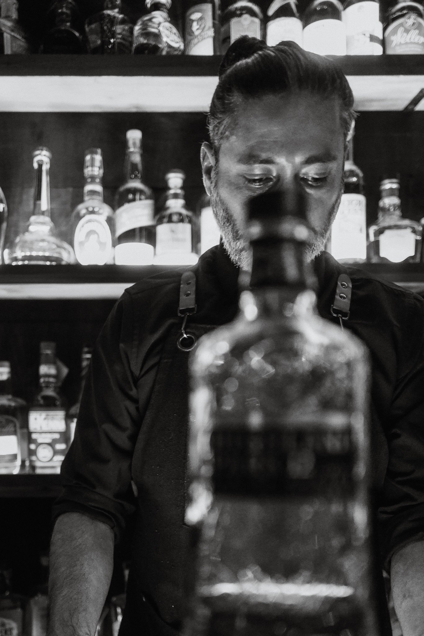 A man with slicked back hair and a beard is looking down at a bottle he is holding in both hands in a store aisle with shelves of bottled beverages in the background. The photo is in black and white.