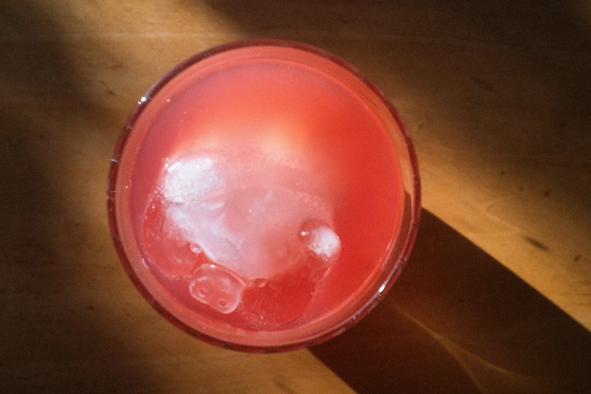 A glass of pink-colored beverage with ice cubes on a wooden surface.