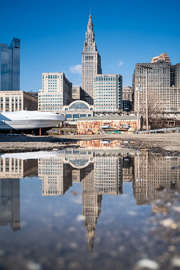 Vertical photo of downtown Cleveland’s Terminal Tower and surrounding office buildings on a clear blue-sky day, reflected sharply in a foreground puddle on rough gravel, creating an upside-down mirror image of the skyline.