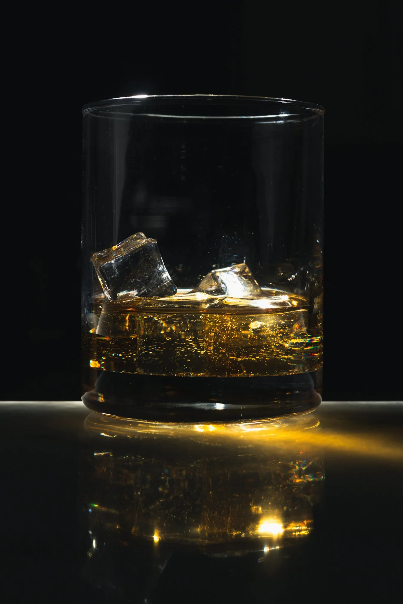A close-up photograph of a clear rocks glass containing amber whiskey over ice cubes, set against a solid black background. Warm golden light highlights the liquid from behind, creating a glowing effect and subtle reflections on the glass and surface