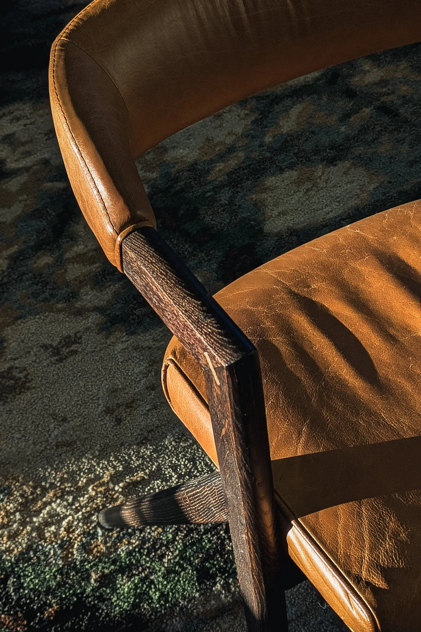 A warm-toned interior scene featuring soft sunlight streaming across a textured beige wall and a framed abstract photograph, followed by a close-up of a brown leather chair with dark wooden arms resting on a patterned rug. The afternoon light emphasi