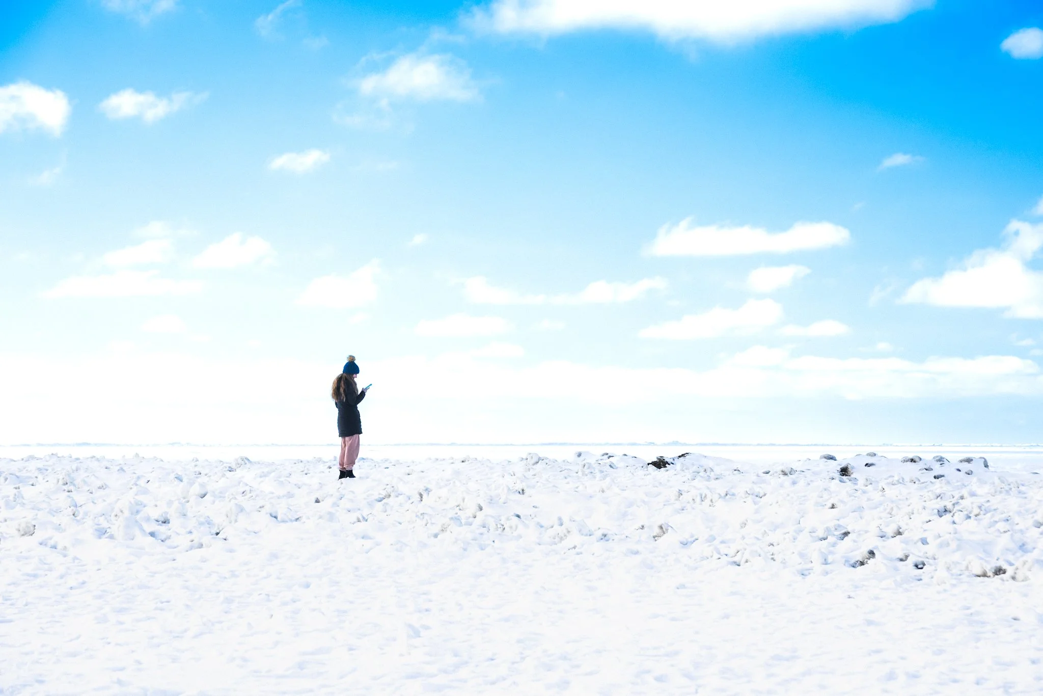A person in winter clothing standing alone in a wide, snow‑covered landscape under a bright blue sky with scattered clouds. The individual wears a dark coat, pink pants, boots, and a blue hat while looking at a handheld device. The snowy outdoor scen