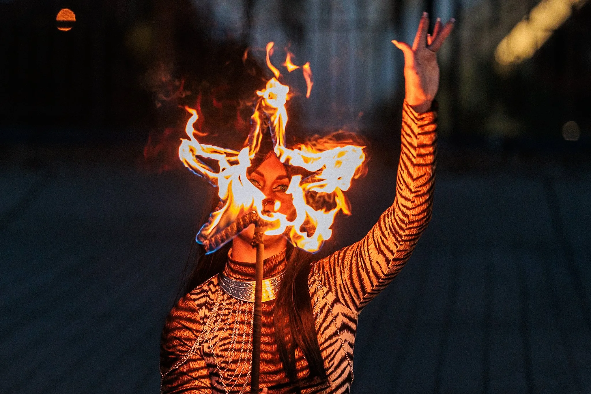 A woman performing with fire, holding a flaming torch and with flames surrounding her face, wearing a tiger-striped outfit in a dark setting.