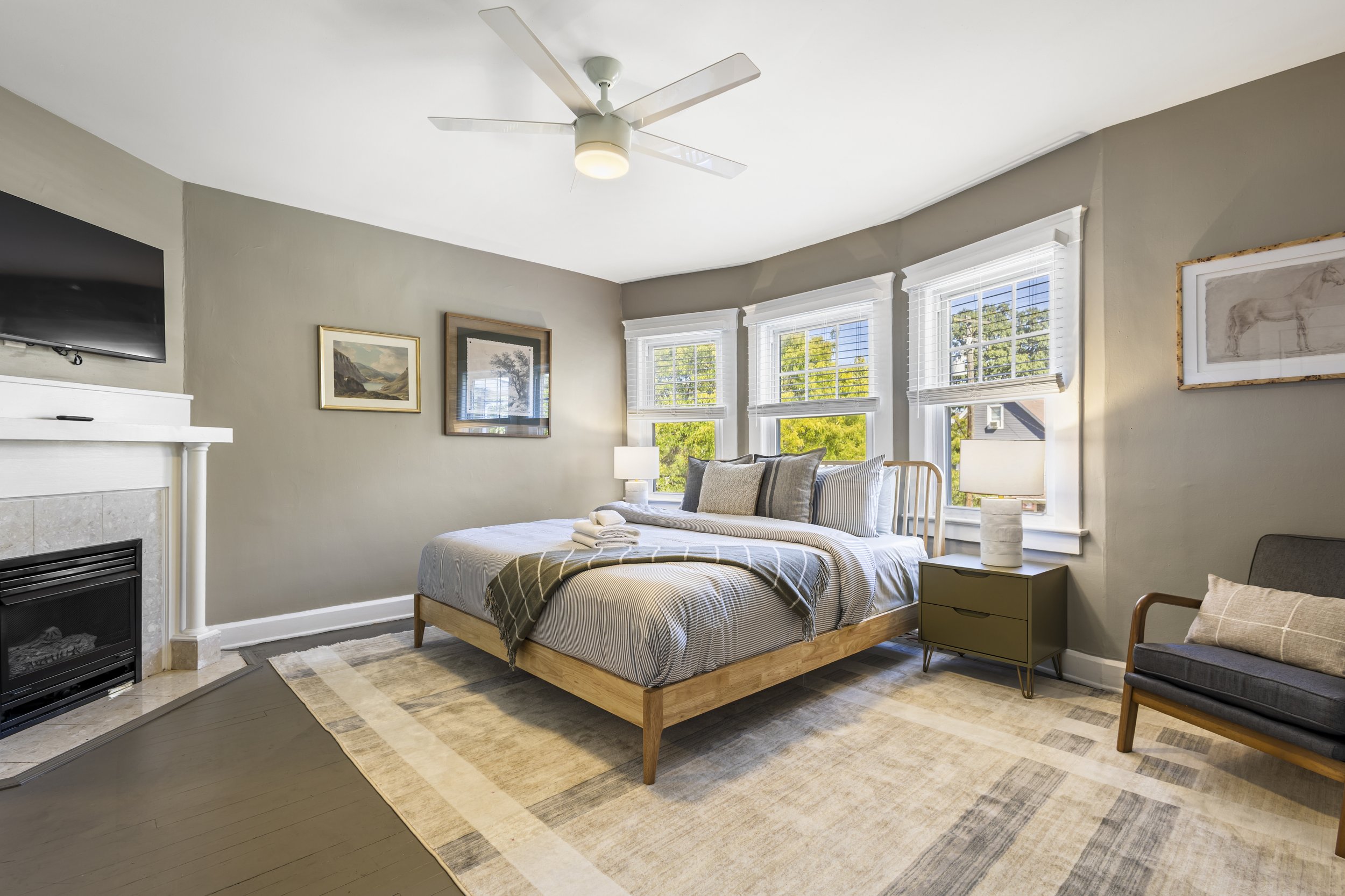 A bedroom with a bed that has striped bedding and pillows, a nightstand with a lamp, a chair with a cushion, three large windows with blinds, paintings on the wall, a ceiling fan, and a rug on wooden flooring.