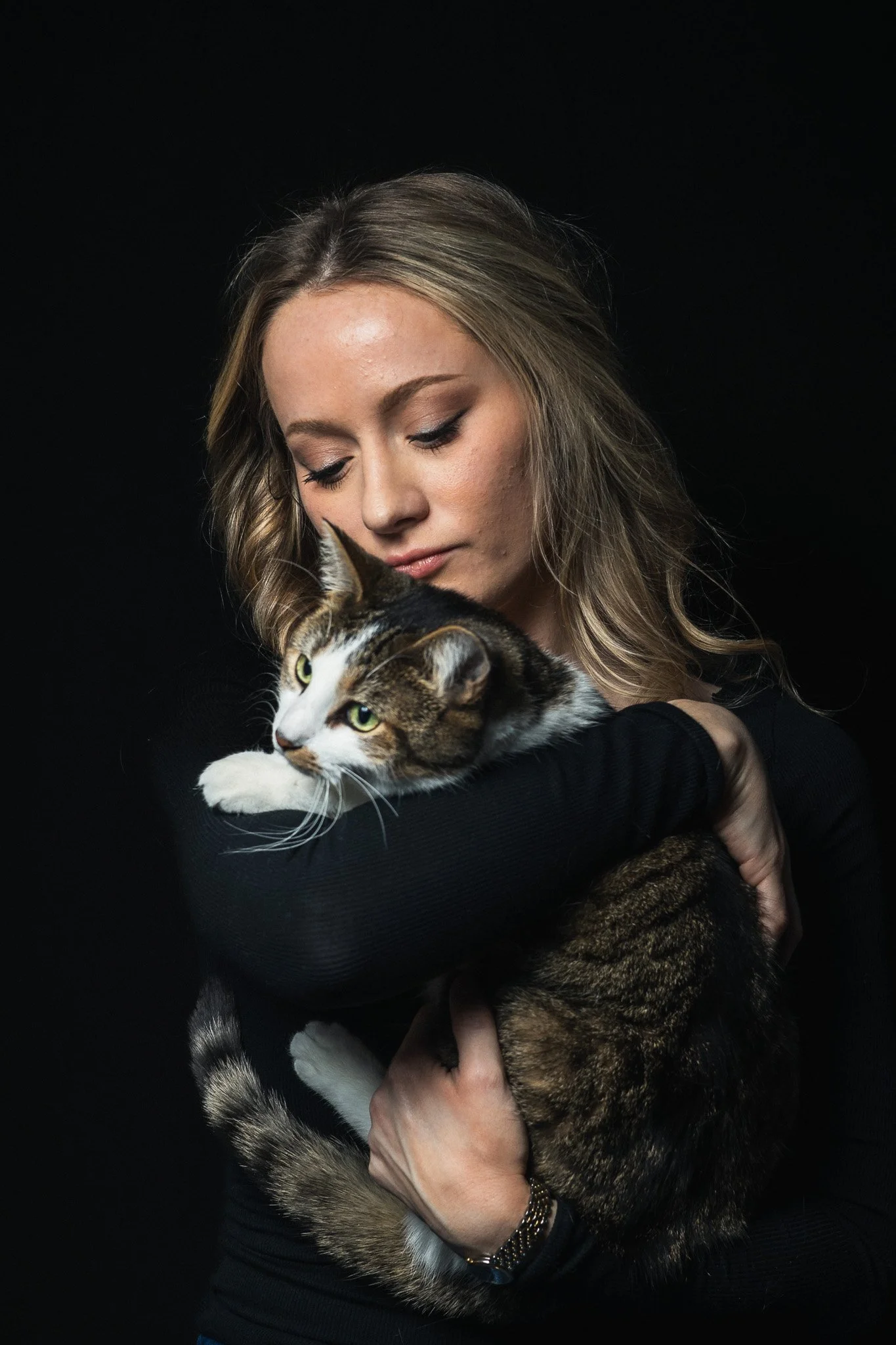 A young woman with long blonde hair, wearing a black long-sleeve shirt, gently holds a tabby and white cat against her chest. The photo is shot against a dark background, highlighting the soft light on both the woman’s face and the cat.