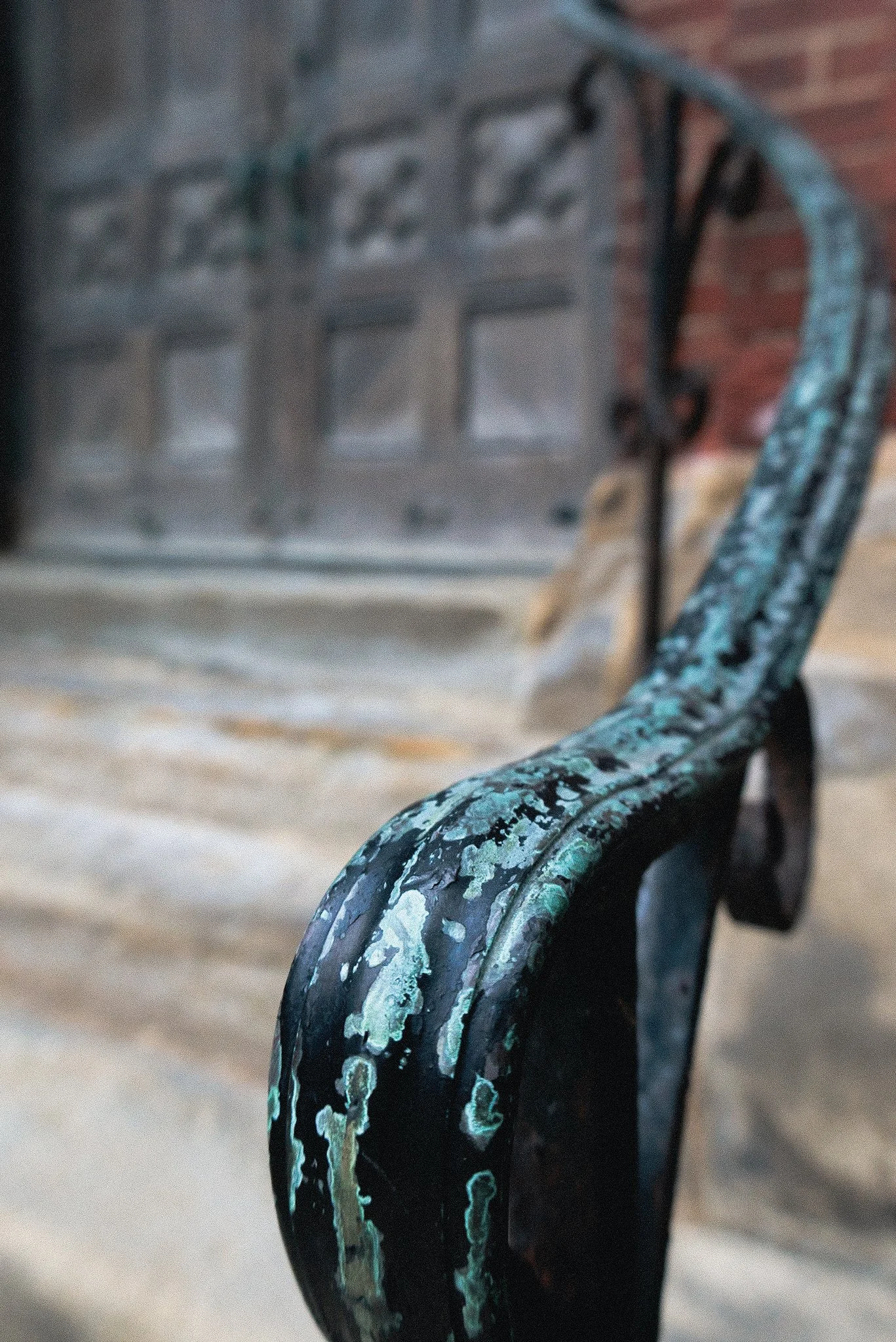 Close-up of a worn, weathered black metal bench with peeling turquoise paint, set against a blurred background of a brick wall and a window with decorative ironwork.