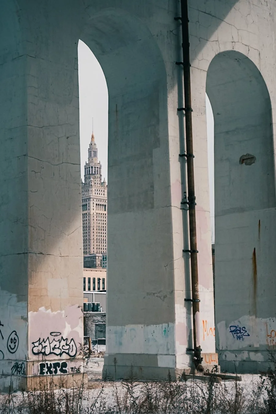 A distant art deco skyscraper Terminal Tower is framed between the massive concrete arches of an overpass, with graffiti at the base of the pillars and a light dusting of snow on the ground.