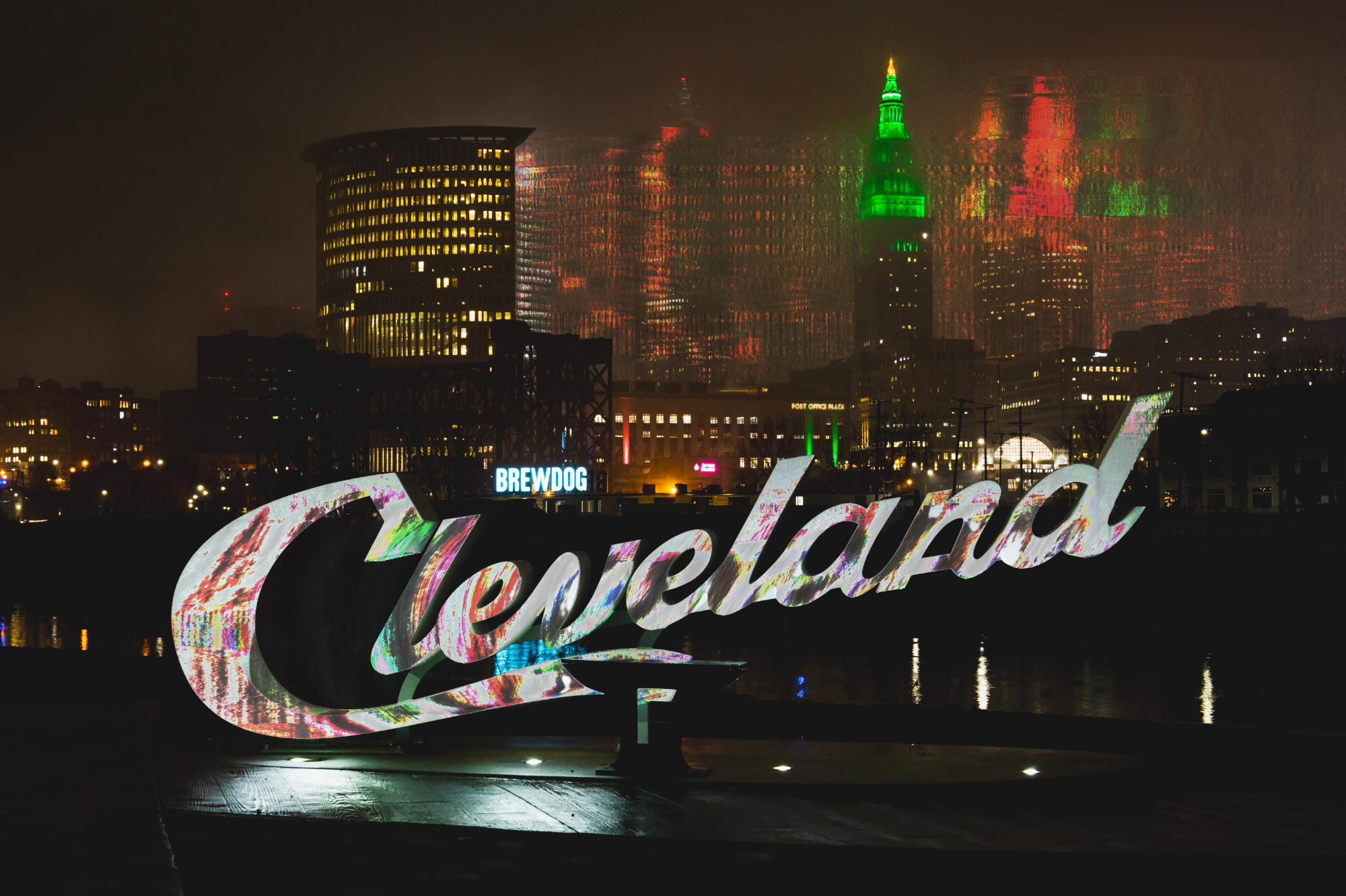 Nighttime view of the Toronto skyline with the iconic 'Toronto' sign illuminated in the foreground, and the city buildings and CN Tower decorated with vibrant lights in the background.