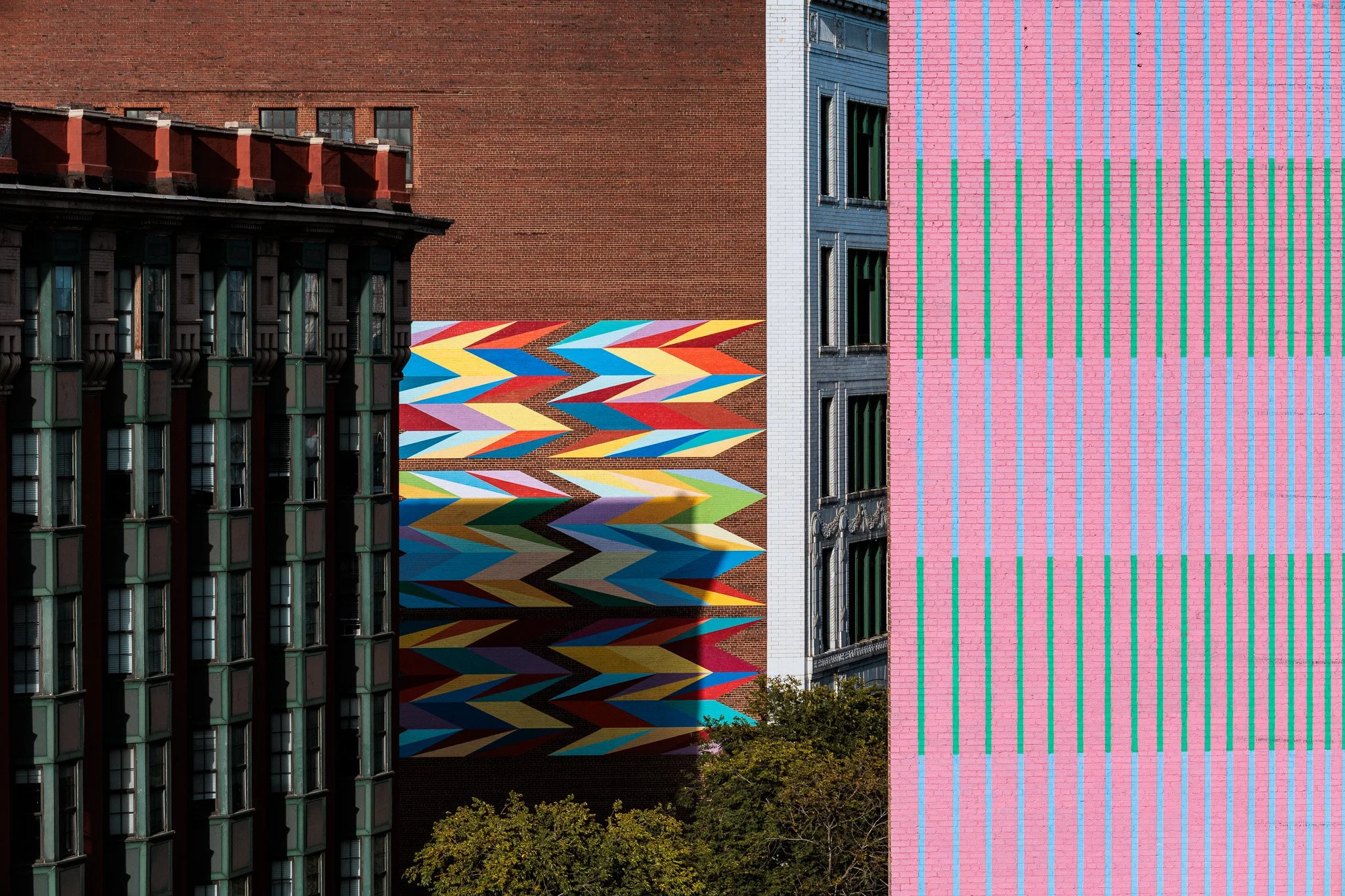 Colorful mural painted on the sides of multiple buildings, featuring bold geometric patterns, stripes, and zigzag designs in pink, blue, yellow, red,, orange, black, and white, with some greenery at the bottom.
