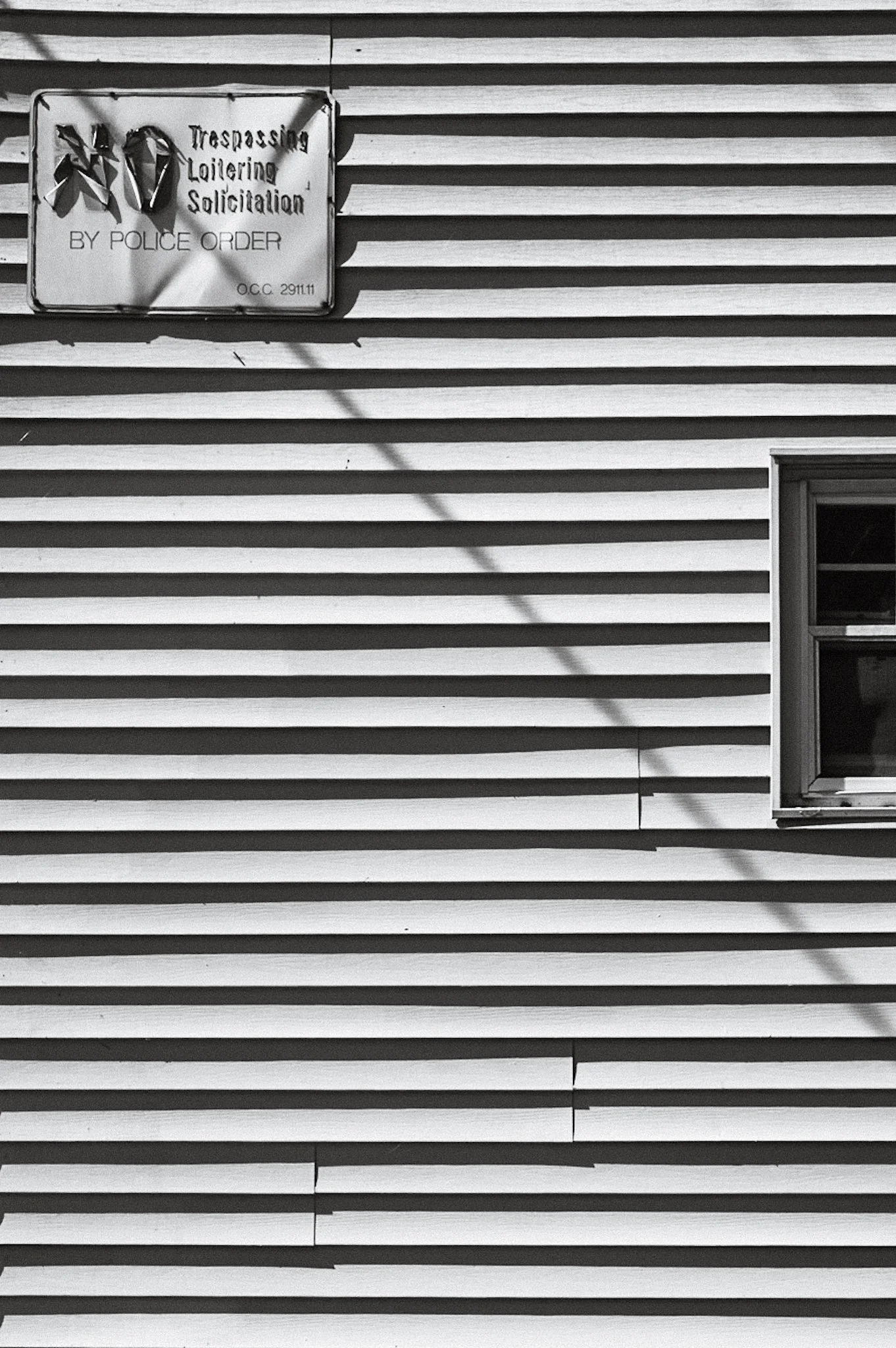 A house with horizontal siding and a window. A warning sign about trespassing, loitering, and solicitation from the police is attached to the house.
