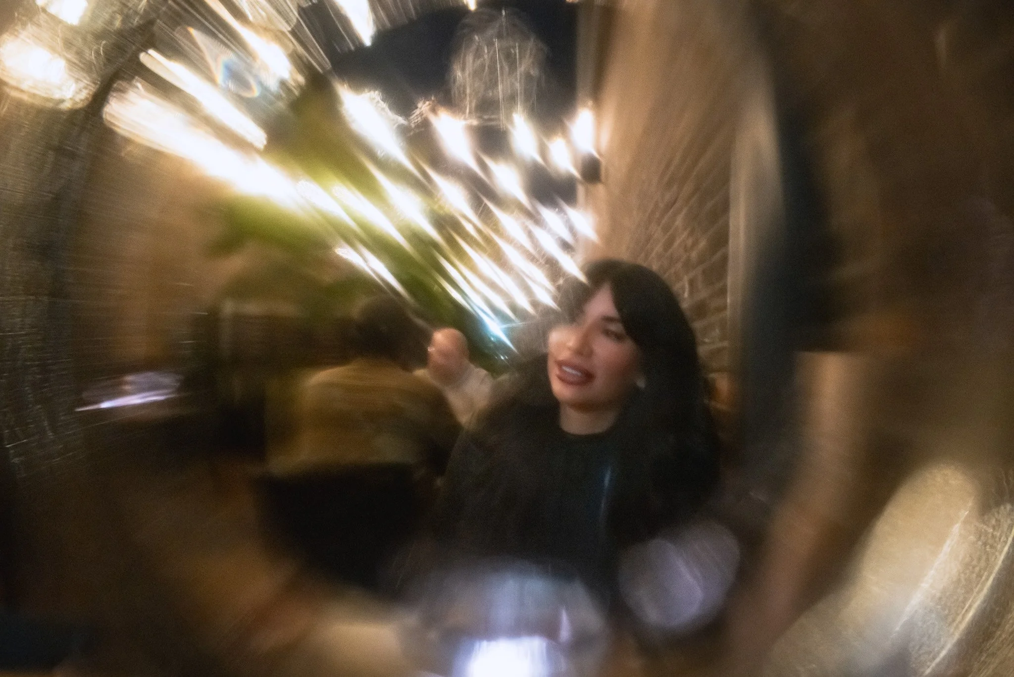 A woman with dark hair and makeup looking through a reflective, metallic surface with blurred lights and other people in the background.